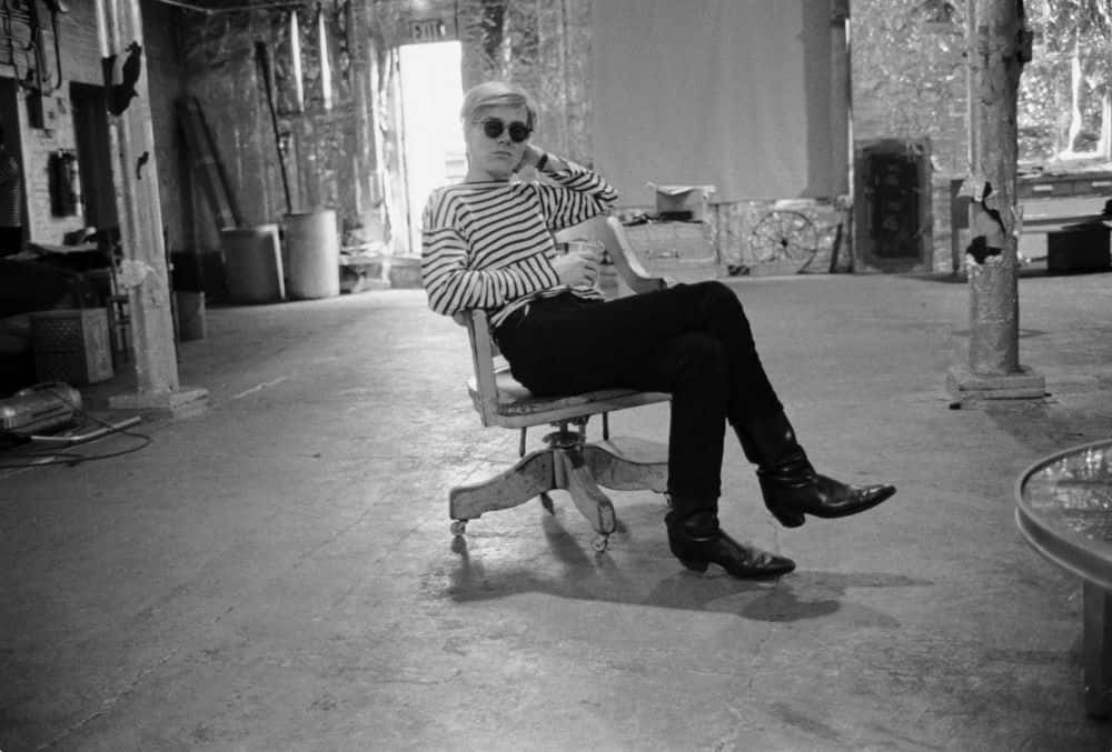 andy warhol 桌布 壁紙 — 一名身穿條紋襯衫和太陽眼鏡的男子，自信地靠在一張有輪子的椅子上，身處一個雜亂且陽光充足的攝影棚空間。