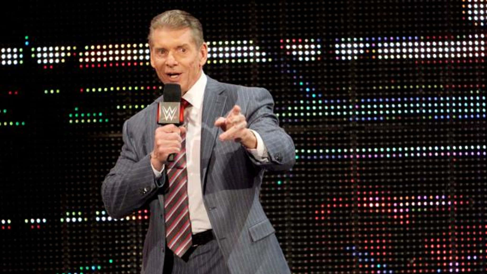 WWE 桌布 壁紙 — 一名身穿條紋西裝的男子拿著WWE麥克風，在充滿活力的LED螢幕背景下用力地比手勢。