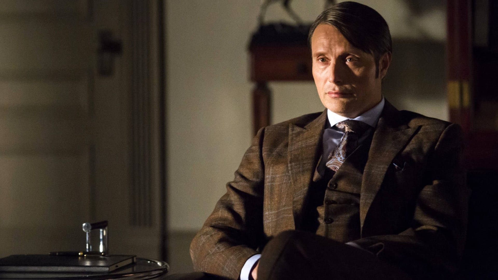 Mads Mikkelsen 桌布 壁紙 — 一名身穿棕色格紋西裝的男子坐在燈光昏暗的房間裡，表情嚴肅，旁邊的桌上放著一個玻璃杯。