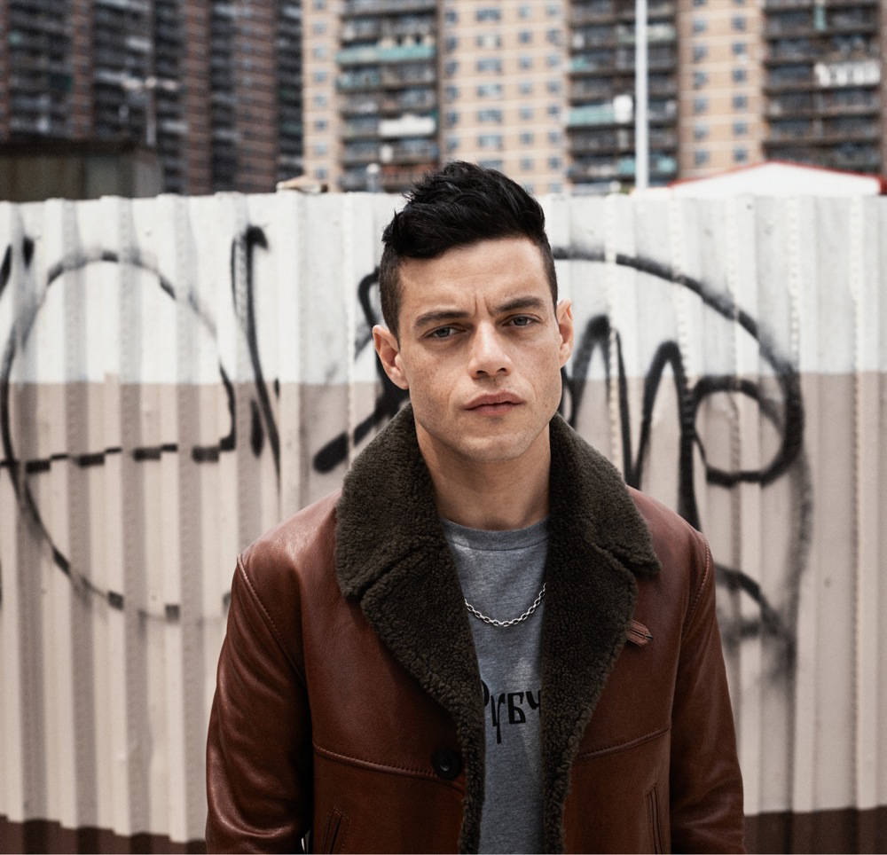 Rami Malek 桌布 壁紙 — 一名身穿棕色皮夾克的年輕男子站在佈滿塗鴉的金屬圍欄前，背後是城市建築。