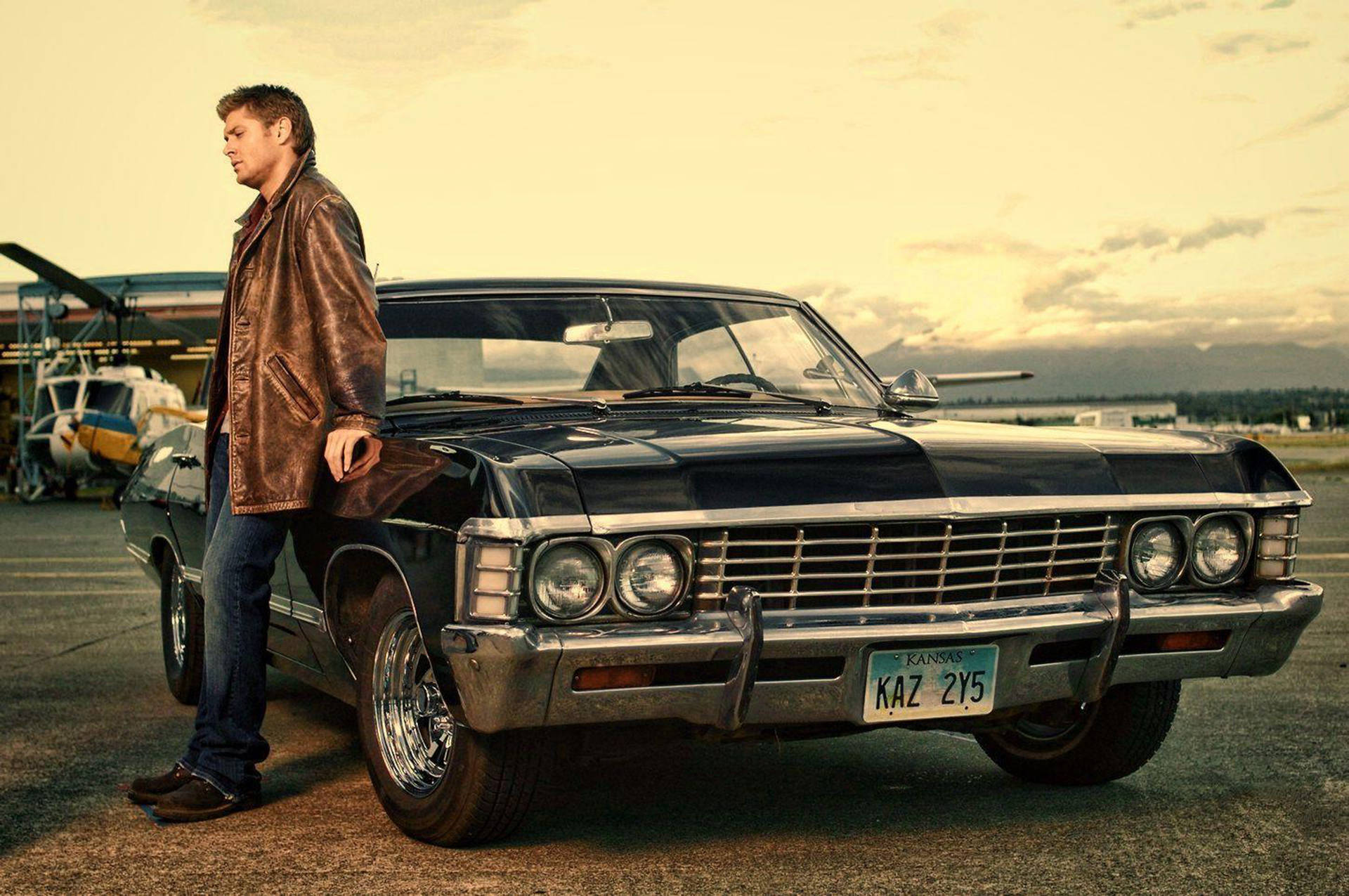 chevrolet impala 1967 桌布 壁紙 — 一名身穿棕色皮夾克的男子在黃金時段的機場停機坪上，靠在一輛帶有鍍鉻細節的黑色經典汽車上。