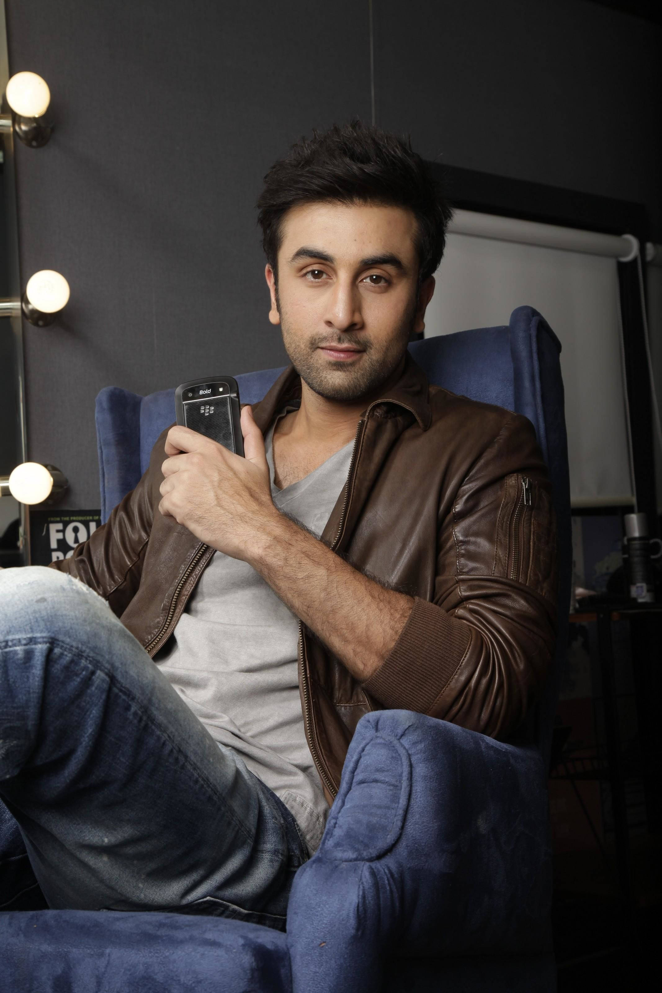 Ranbir Kapoor 桌布 壁紙 — 一名身穿棕色皮夾克的男子放鬆地坐在藍色扶手椅上，手持電話，身後有攝影棚燈光照射。