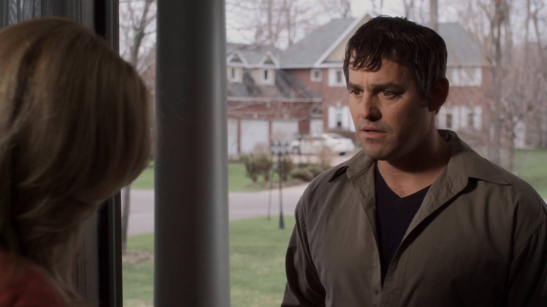 Nicholas Brendon 桌布 壁紙 — 一名身穿棕色襯衫的男子站在窗邊，面對著一位金髮女子，位於郊區住宅內，窗外可見光禿的樹木和磚房。