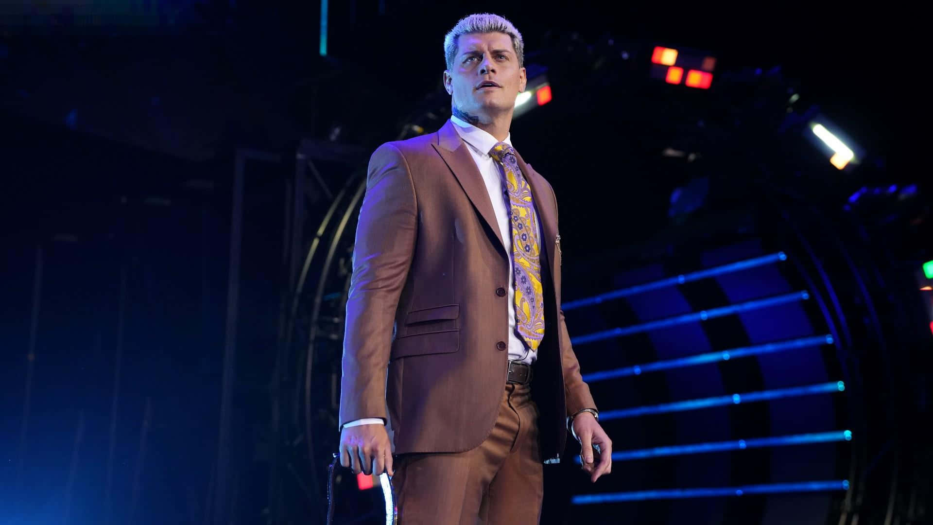 WWE 桌布 壁紙 — 一名身穿棕色西裝和圖案領帶的摔角手自信地站在一個有藍色燈光和黑暗背景的明亮舞台上。