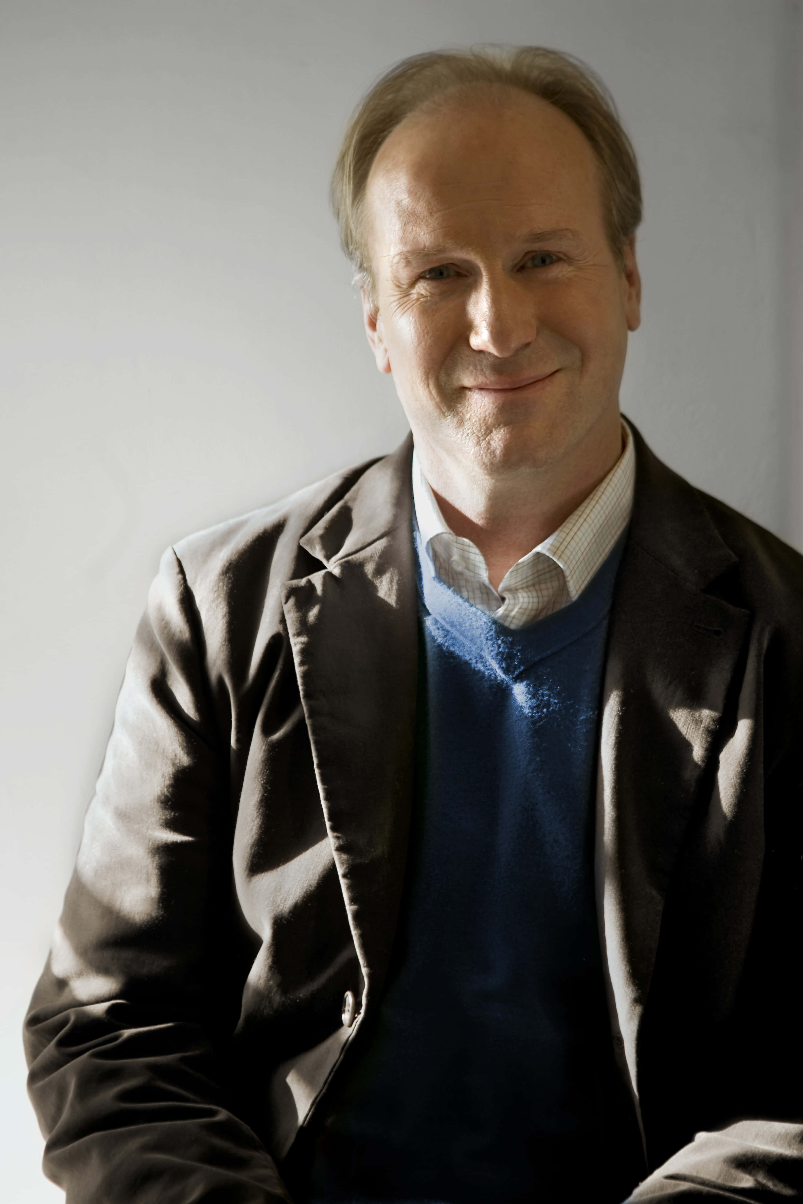 William Hurt 桌布 壁紙 — 一名身穿棕色西裝外套和藍色毛衣的男子在純灰色背景前溫暖地微笑。