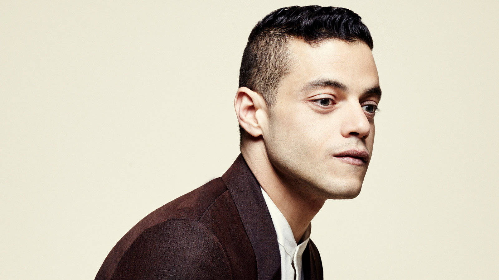 Rami Malek 桌布 壁紙 — 一名身穿棕色西裝外套的男子，在柔和的米色背景前若有所思地凝視，微妙的燈光突顯他銳利的五官。