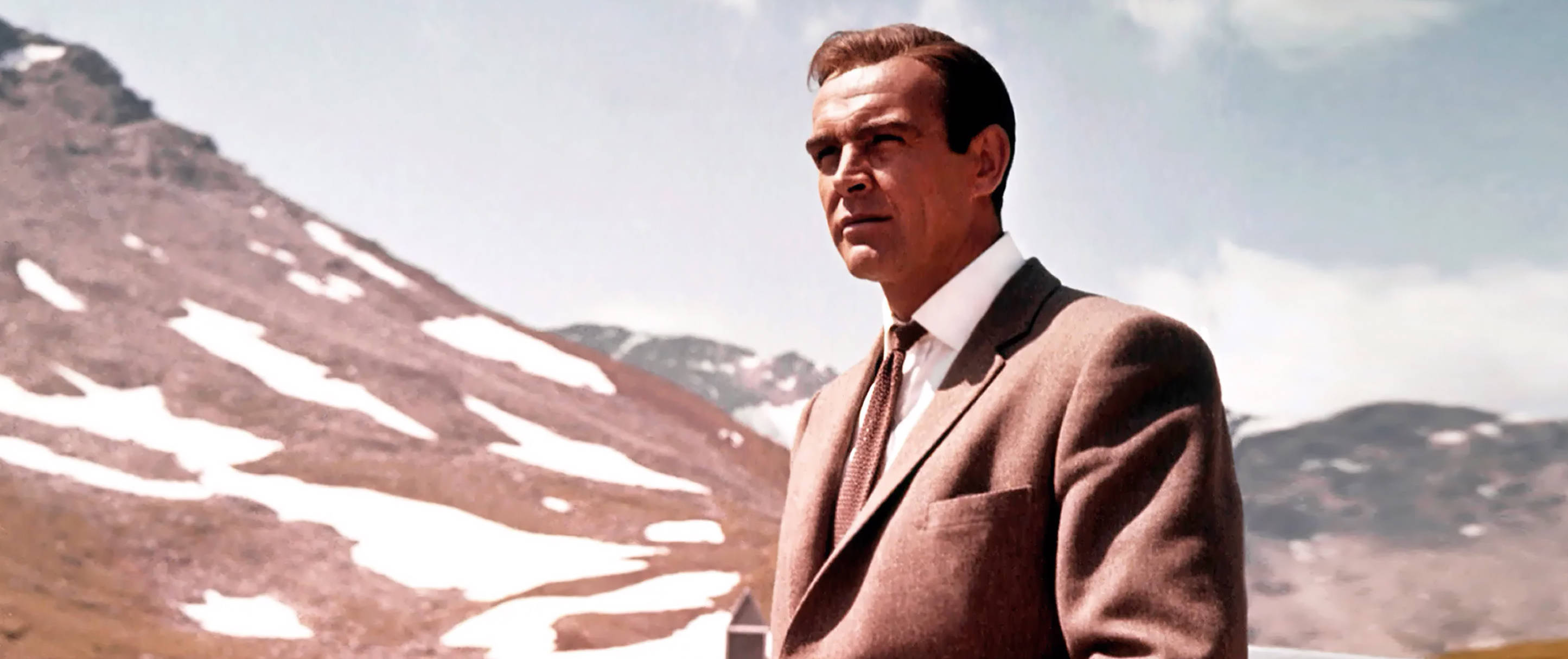 Sean Connery 桌布 壁紙 — 一名身穿棕色西裝的男子在明亮的天空下，自信地站在被雪覆蓋的山脈背景下。