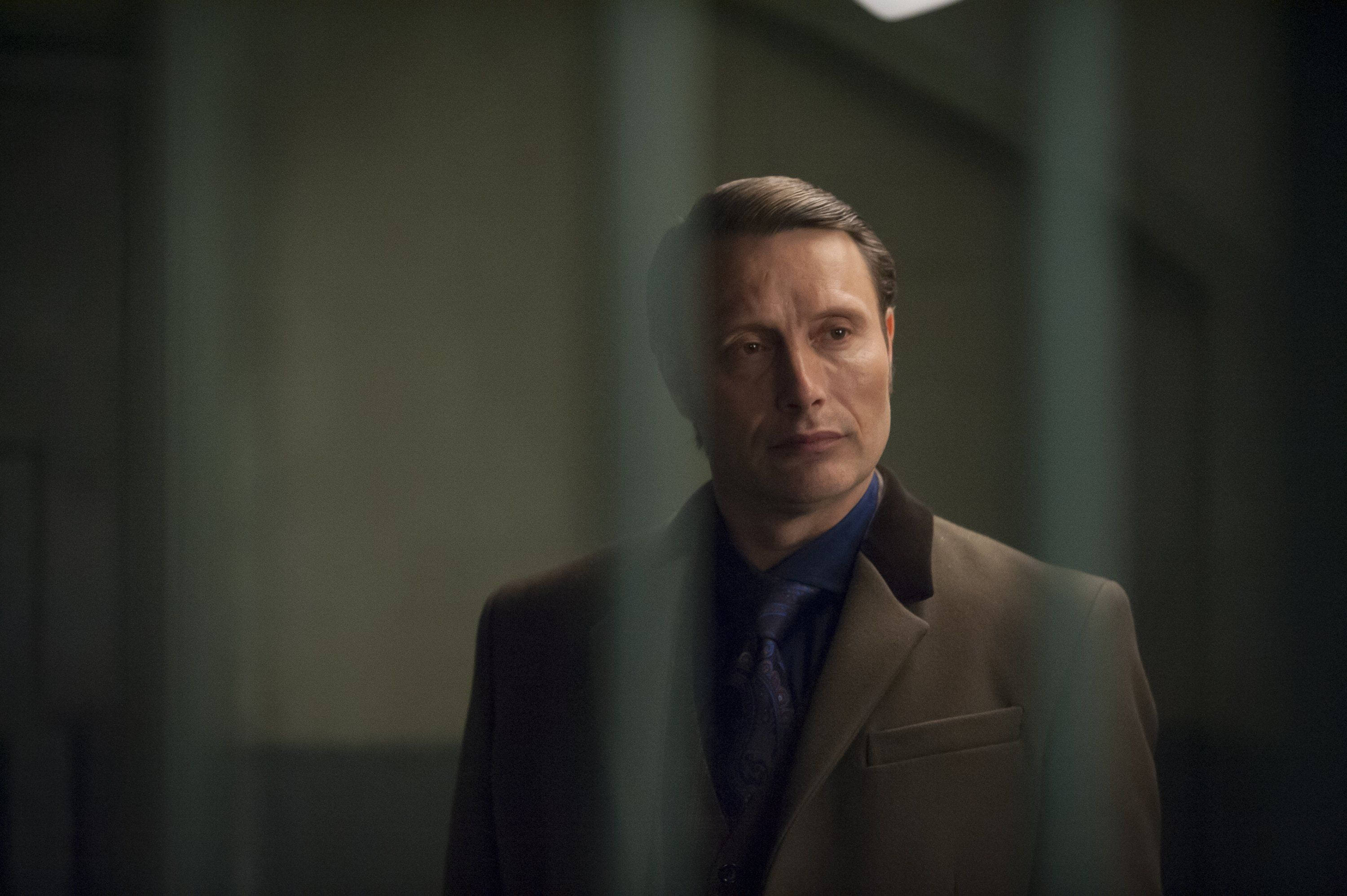 Mads Mikkelsen 桌布 壁紙 — 一名身穿棕色西裝的男子站在昏暗房間的垂直鐵欄後，表情嚴肅。