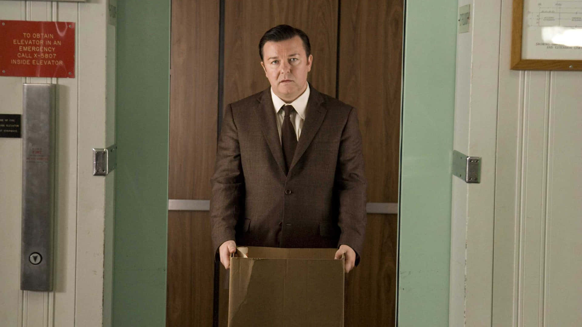 ricky gervais 桌布 壁紙 — 一名身穿棕色西裝的男子站在電梯大廳，手持紙箱，表情嚴肅。