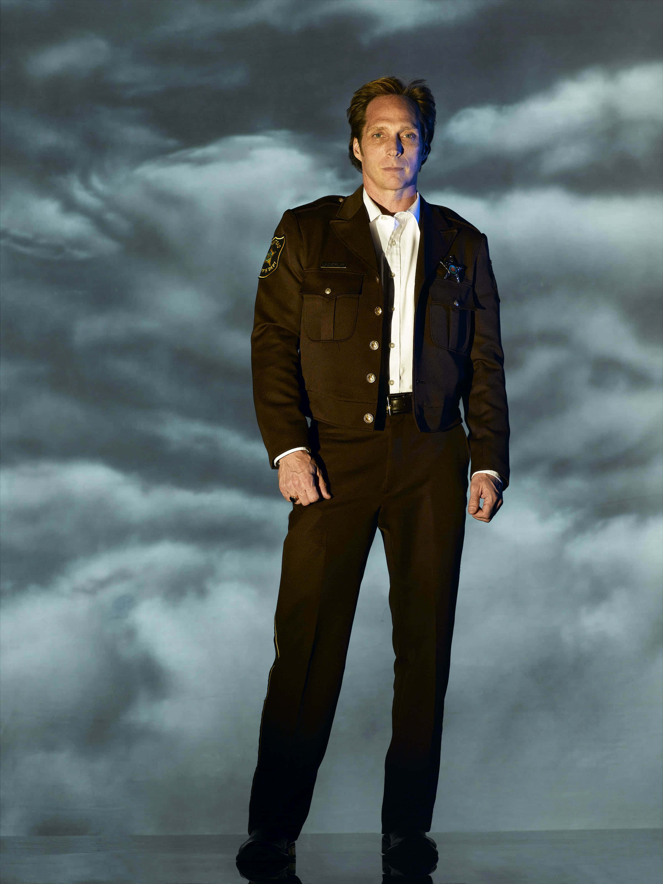 William Fichtner 桌布 壁紙 — 一名身穿棕色警長制服的男人，在戲劇性的多雲天空背景下莊嚴地站立。