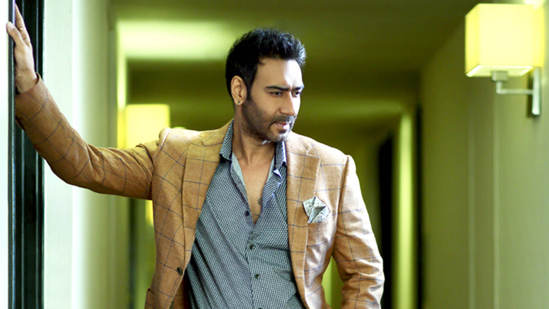 ajay devgn 桌布 壁紙 — 一名身穿棕褐色格紋西裝外套的男子在一個溫暖照明的走廊中靠著牆，周圍有黃色的環境燈光。