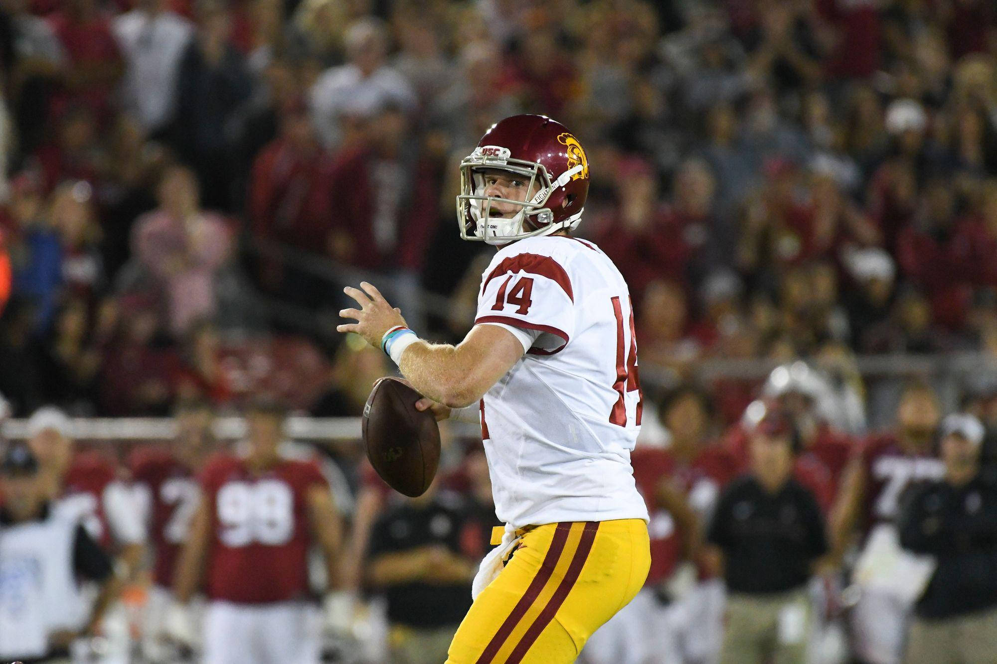 Football Players 桌布 壁紙 — 一名身穿棗紅色和金色球衣的美式足球四分衛在球場燈光下投球，背景是模糊的球迷。