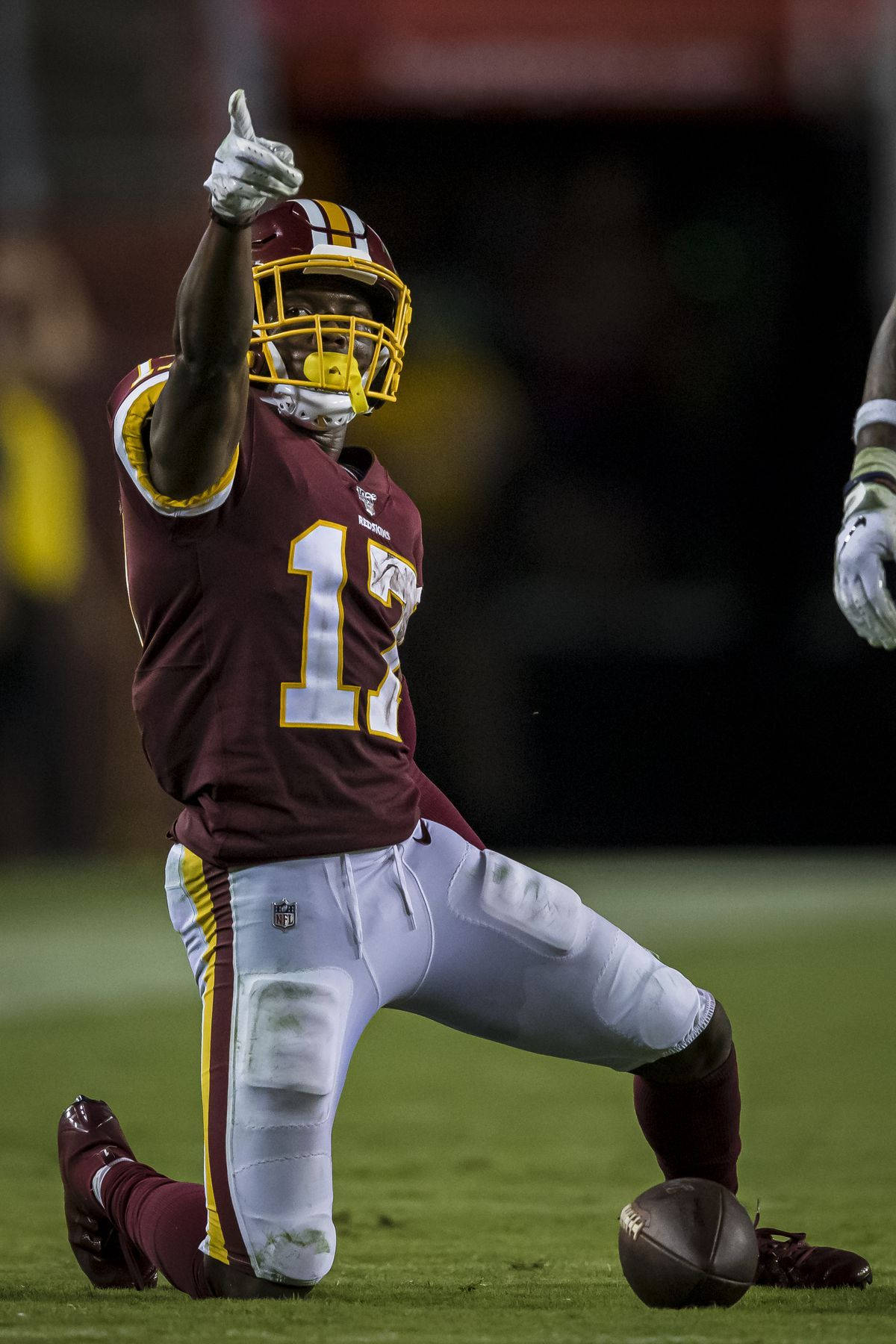 Football Players 桌布 壁紙 — 一名身穿棗紅色和金色球衣的美式足球員在球場燈光下跪在草地上，豎起大拇指，腳邊放著一個足球。