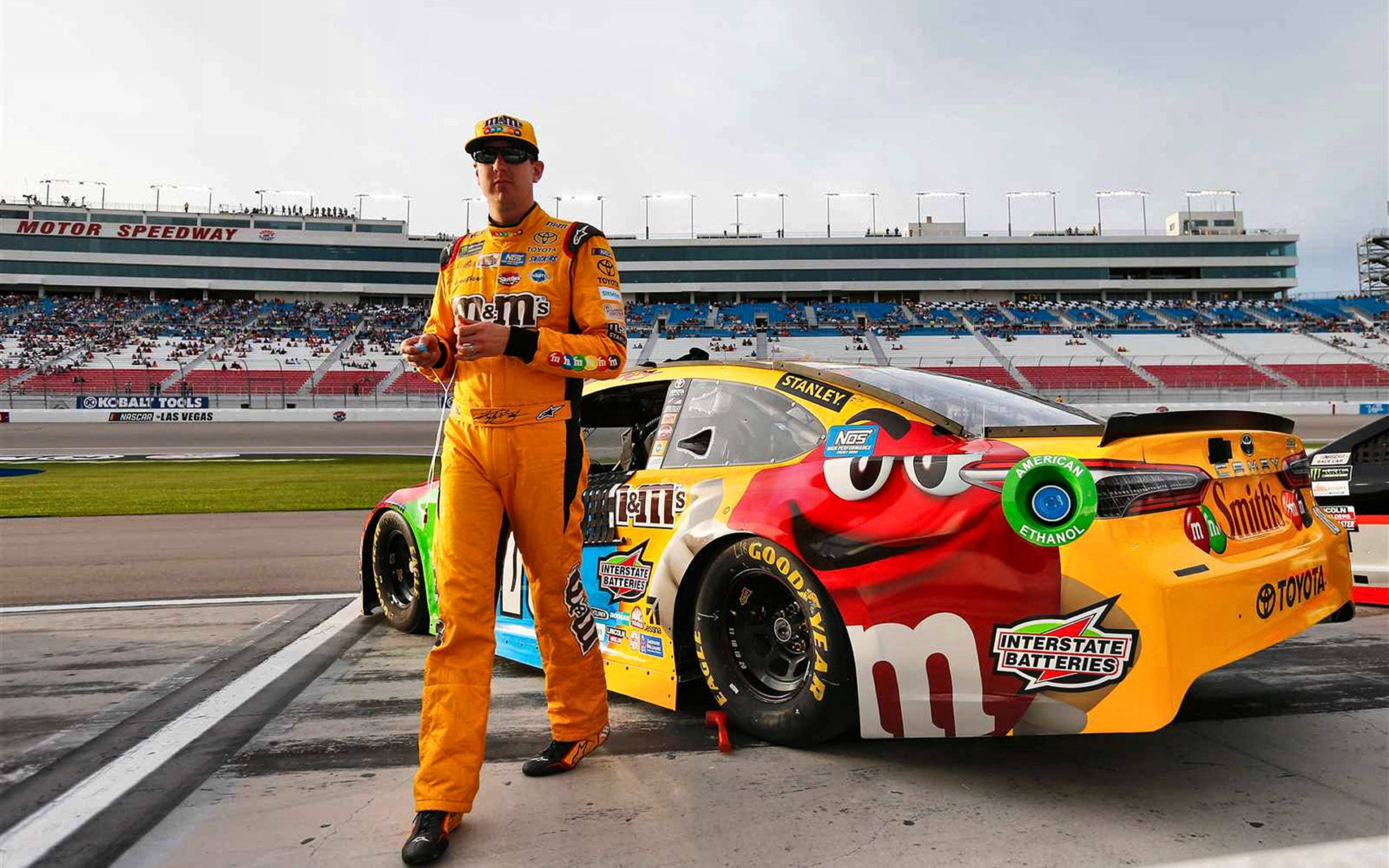 Racing 桌布 壁紙 — 一名身穿橙色M&M's賽車服的NASCAR車手站在擁擠的賽車道上，旁邊是他色彩繽紛的M&M's主題Toyota賽車。