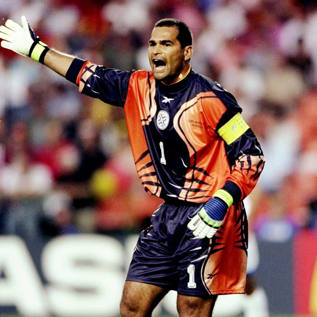 足球 桌布 壁紙 — 一名身穿橙色和海軍藍球衣的足球守門員在球場燈光下比賽時伸出手臂大喊。