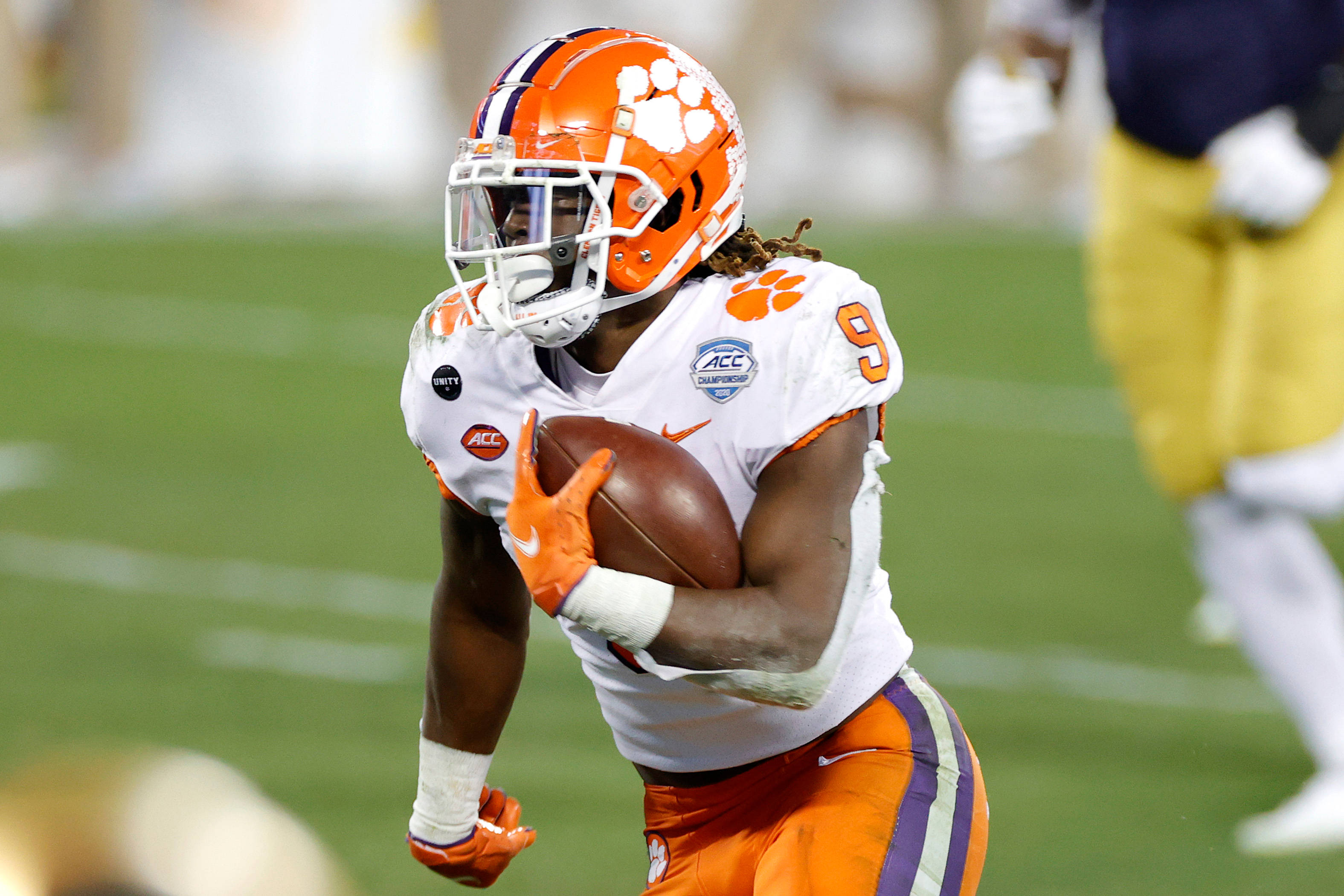 Travis Etienne 桌布 壁紙 — 一名身穿橙色和白色裝備的克萊姆森美式足球員在體育場燈光下的綠色場地上帶球奔跑。