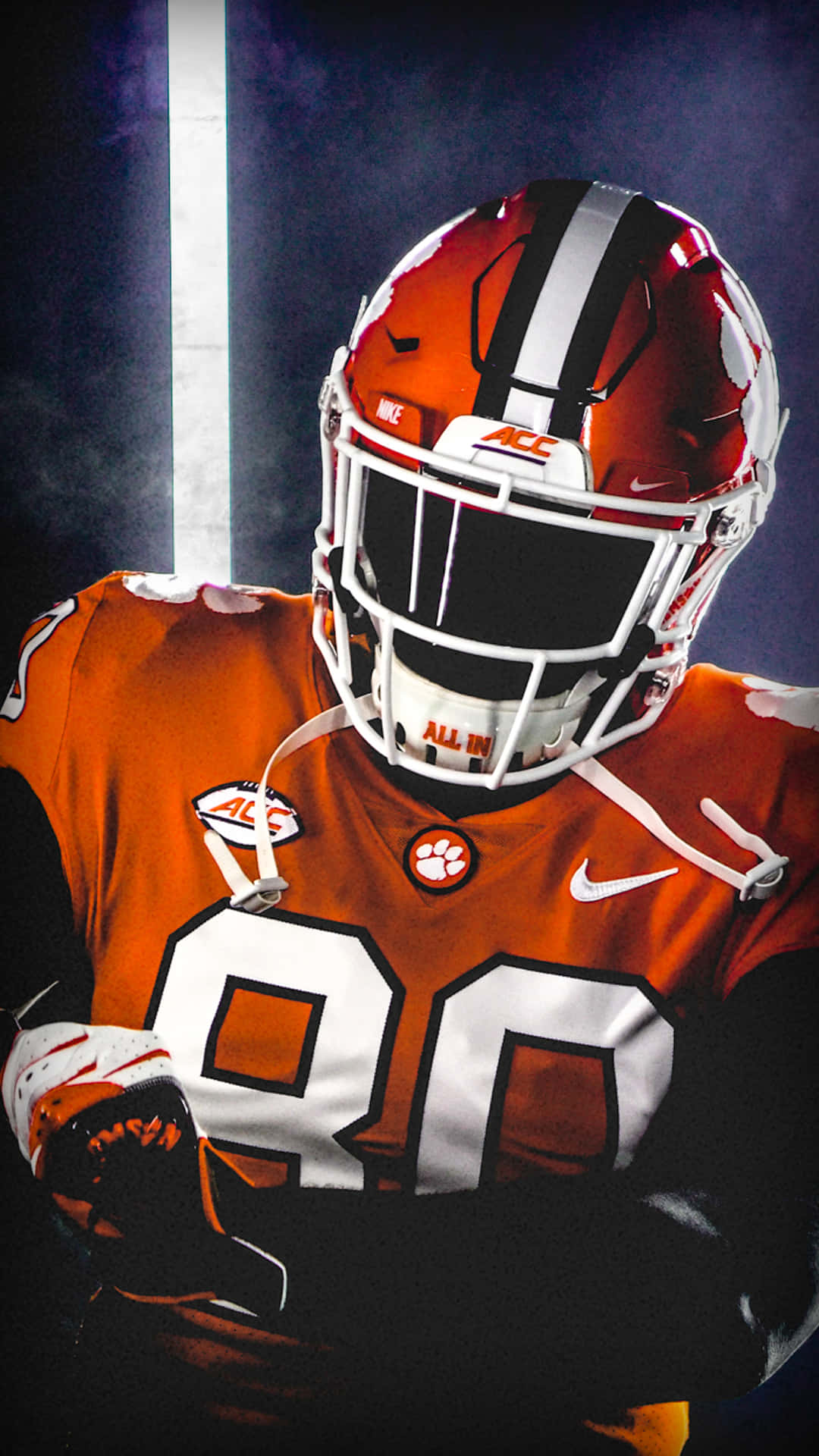 Clemson iPhone 桌布 壁紙 — 一名身穿橙色和白色裝備的克萊姆森足球運動員站在戲劇性的聚光燈下，煙霧在他周圍盤旋。