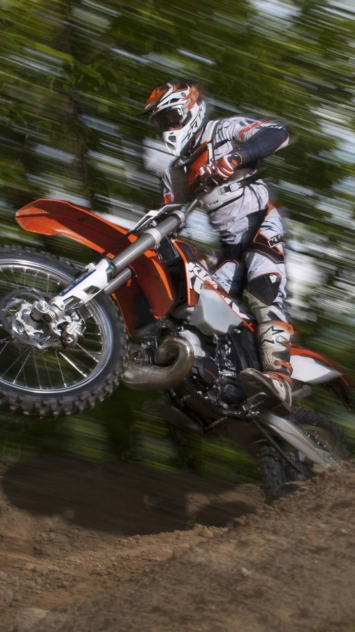 機車 桌布 壁紙 — 一名身穿橙色和白色裝備的越野摩托車手在 dirt bike 上傾斜入彎，穿過一片模糊的綠色樹木。