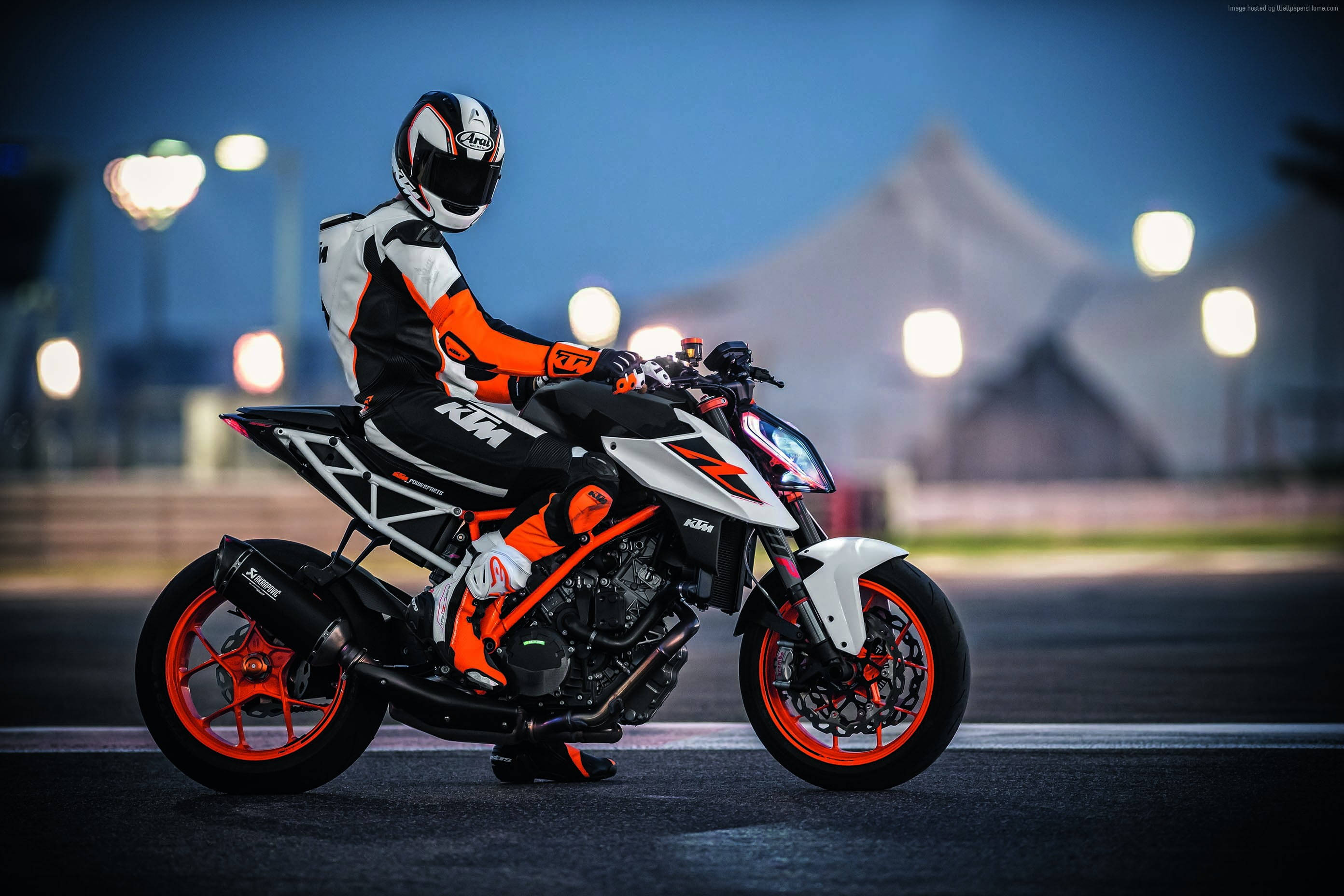 KTM 摩托車 桌布 壁紙 — 一名身穿橙色和白色裝備的車手，在暮色賽道背景下，坐在一輛帶有發光橙色輪圈的KTM摩托車上，蓄勢待發。