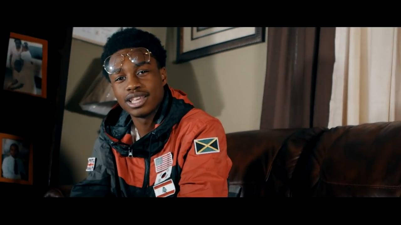 Lil Tjay 桌布 壁紙 — 一名身穿橙色和黑色夾克、帶有旗幟的年輕男子坐在皮革沙發上，房間光線溫暖，牆上掛著裱框照片。