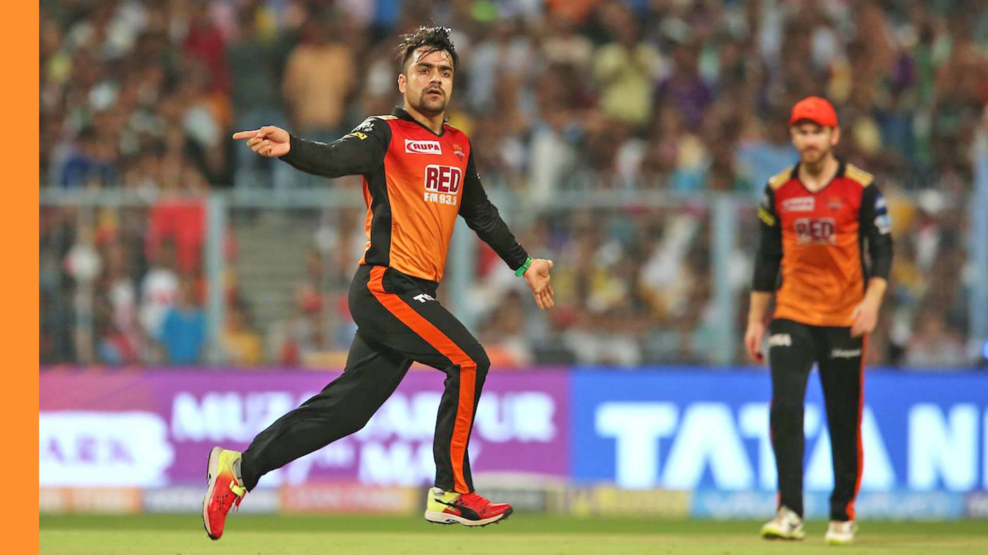 Sunrisers Hyderabad 桌布 壁紙 — 一名身穿橙色和黑色球衣的板球投球手在燈光明亮的球場上動作，背景有模糊的觀眾。