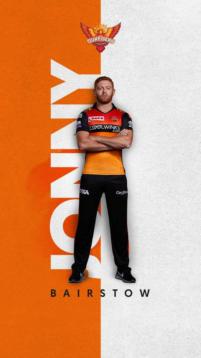 Sunrisers Hyderabad 桌布 壁紙 — 一名身穿橙色和黑色球衣的板球運動員，在鮮明的橙色和白色分割背景前自信地交叉雙臂站立。