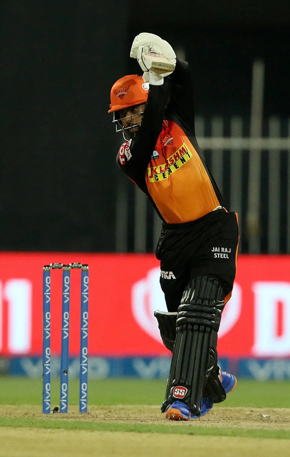 Ipl 2021 桌布 壁紙 — 一名身穿橙色和黑色裝備的板球擊球手在體育場燈光下高舉雙臂慶祝勝利。