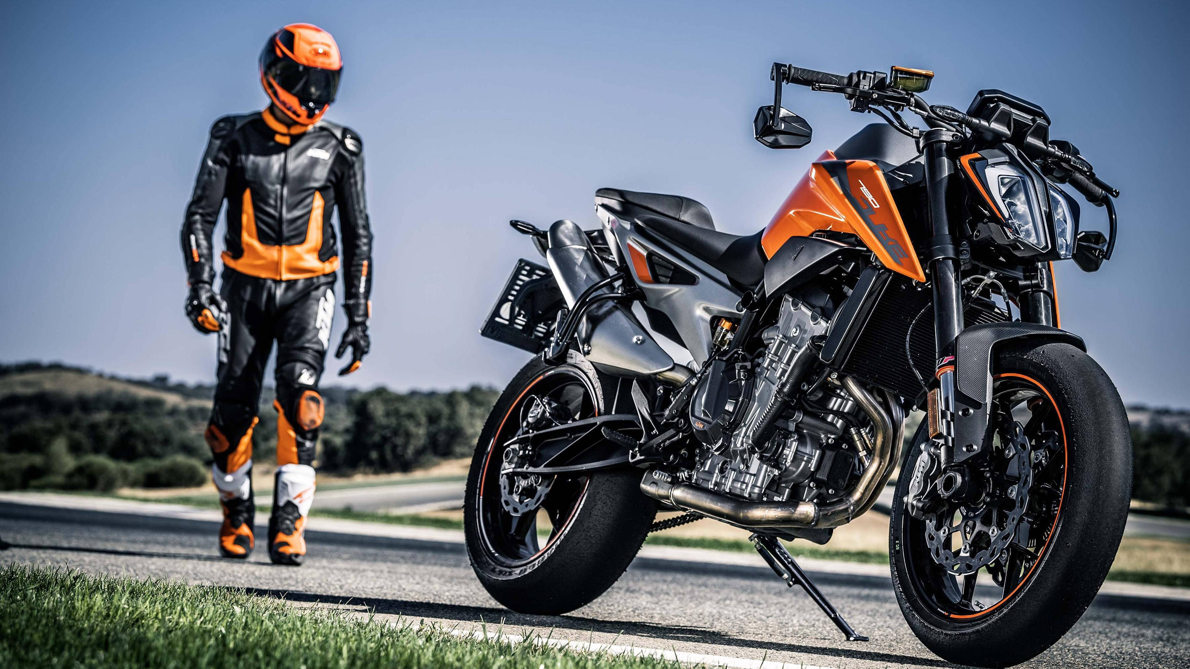 KTM 摩托車 桌布 壁紙 — 一名身穿橙色和黑色裝備的騎士，走在一輛停在陽光普照道路上的時尚橙色和黑色機車旁邊。