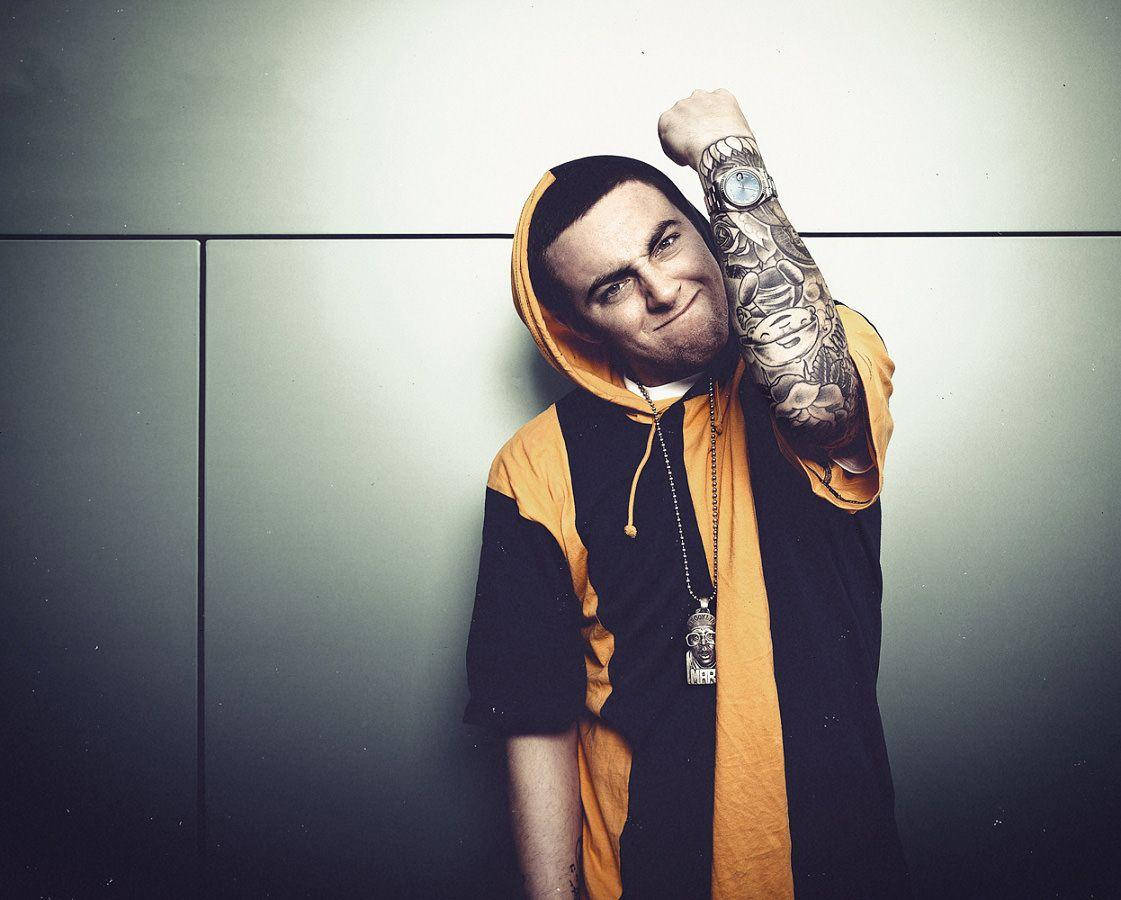 Mac Miller 桌布 壁紙 — 一名身穿橙色和黑色連帽衫的男子，紋身的手臂抬起，戴著骷髏吊墜項鍊，靠著灰色牆壁。