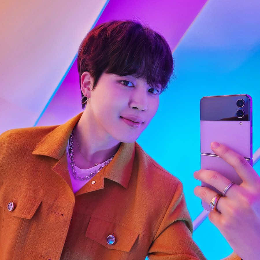 Bts Endorsements 桌布 壁紙 — 一名身穿橙色外套的年輕男子在充滿活力的紫色和藍色幾何背景前微笑自拍。