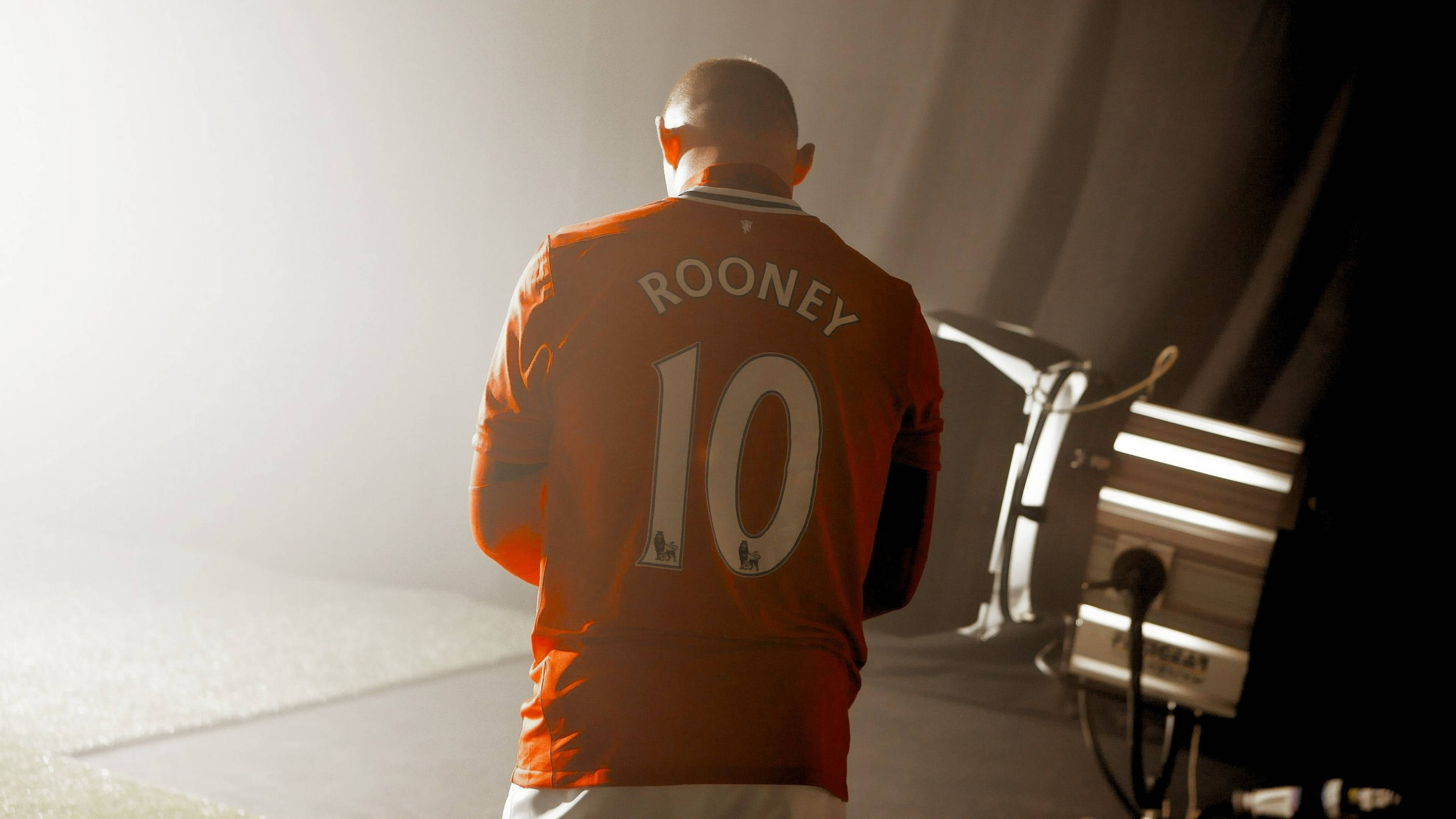 Rooney 桌布 壁紙 — 一名身穿橙色朗尼10號球衣的足球員背對鏡頭，站在由專業燈光照亮的朦朧攝影棚中。