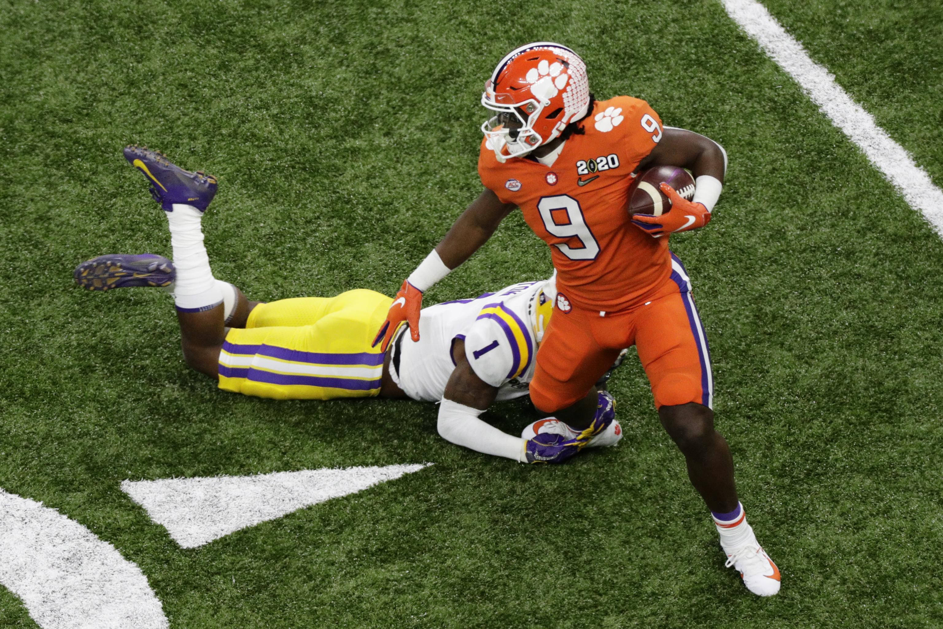Travis Etienne 桌布 壁紙 — 一名身穿橙色球衣的克萊姆森足球員帶球奔跑，而一名身穿黃色球衣的LSU防守球員在綠色草地上擒抱他。