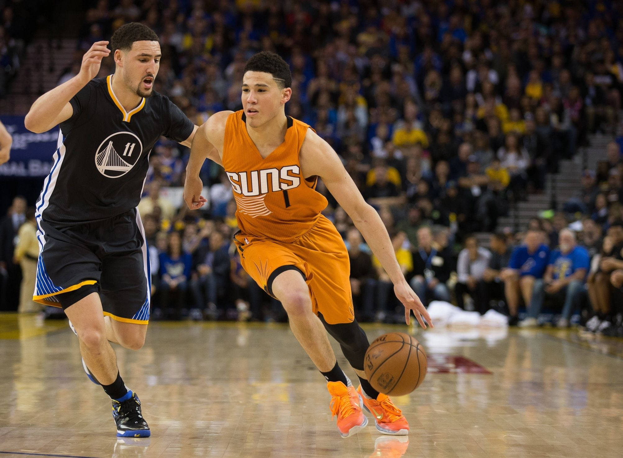 Devin Booker 桌布 壁紙 — 一名身穿橙色的鳳凰城太陽隊球員在滿座的NBA比賽中運球，同時被一名身穿黑色的金州勇士隊球員緊密防守。