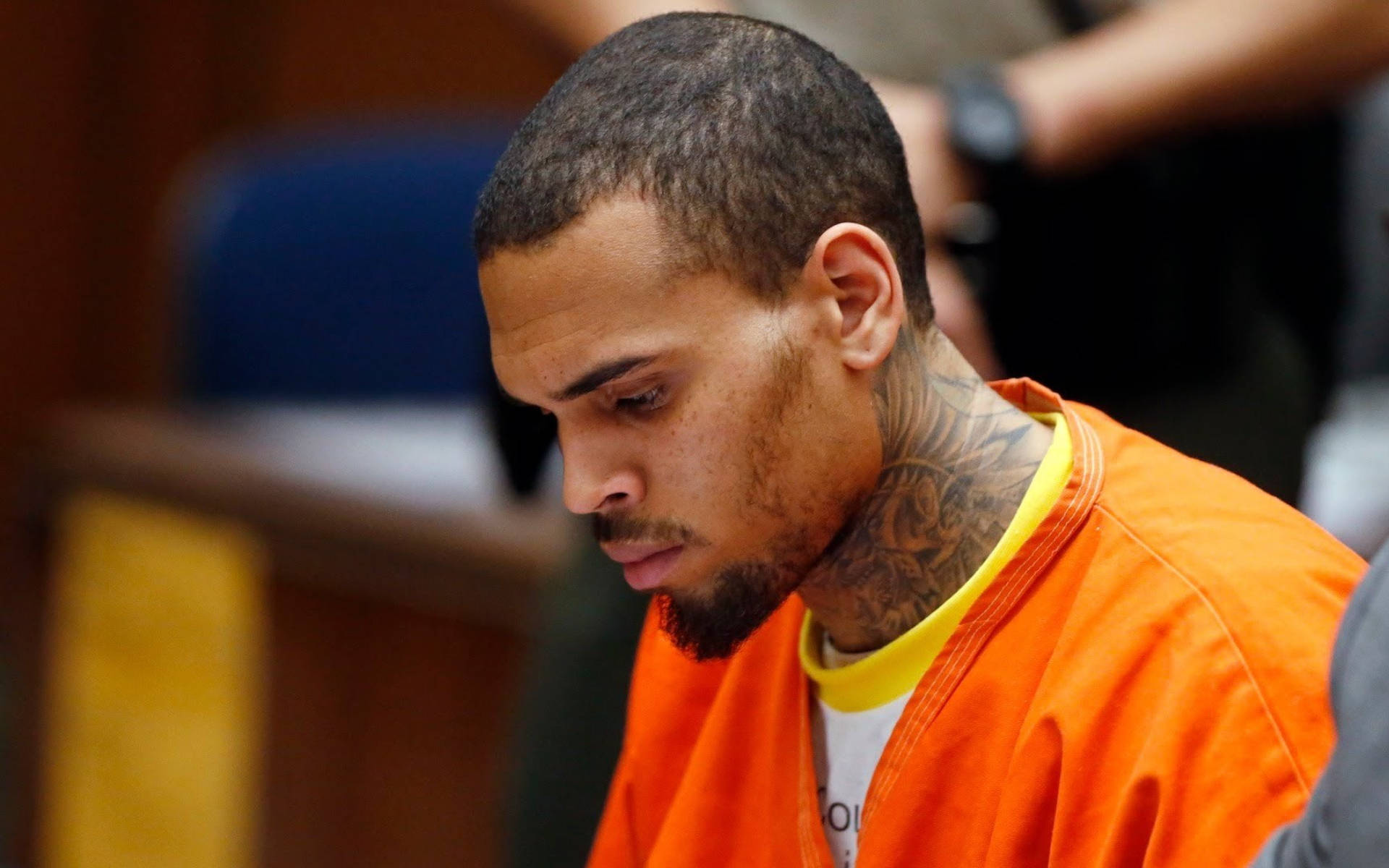 Chris Brown 桌布 壁紙 — 一名身穿橙色監獄連身衣、頸部有明顯刺青的男子在法庭上低頭。