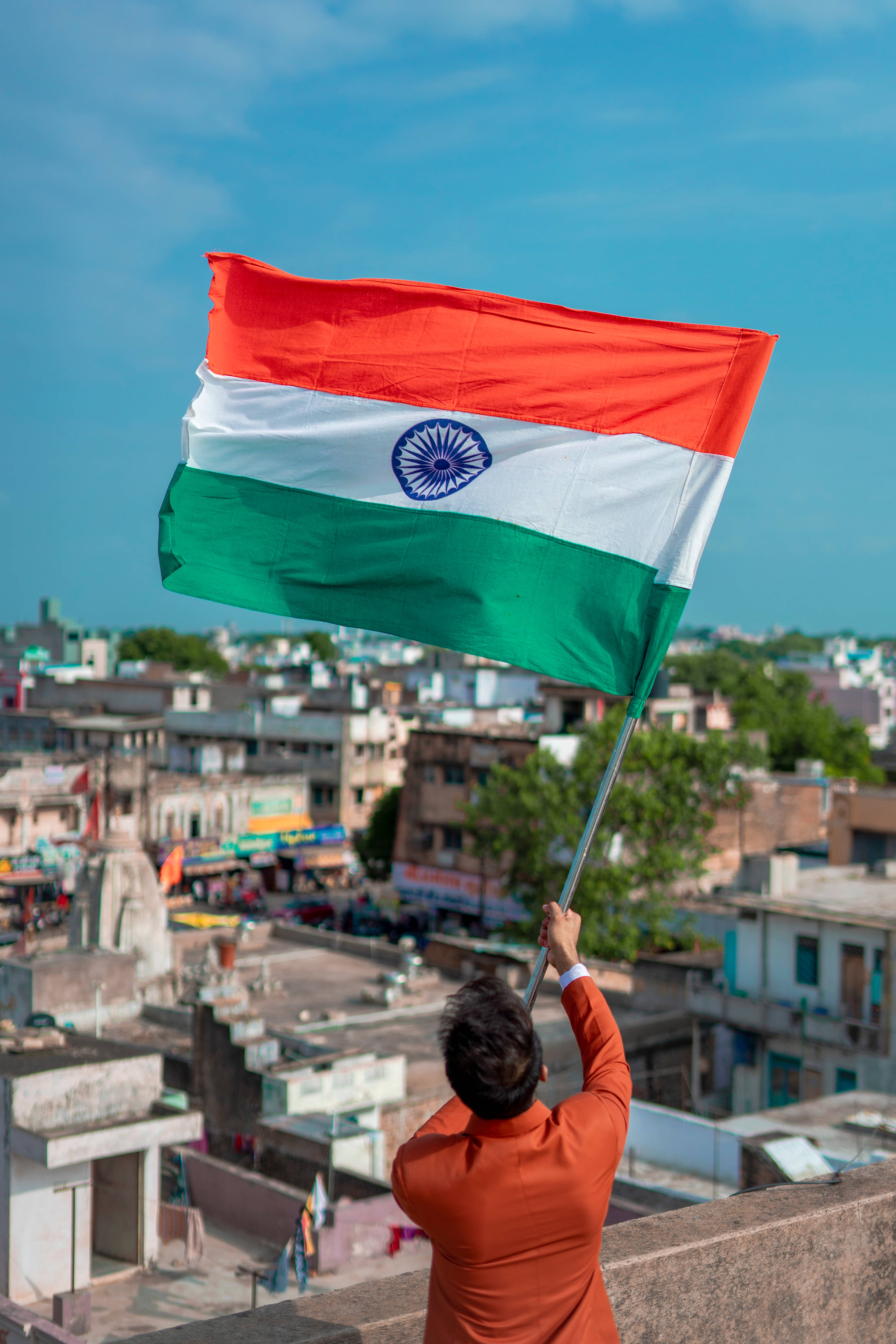 Tiranga 桌布 壁紙 — 一名身穿橙色衣服的人在高樓大廈的天台上，在晴朗的藍天下高舉印度國旗揮舞。