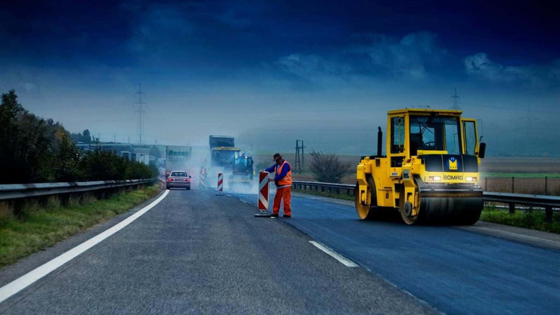 Infrastructure 桌布 壁紙 — 一名身穿橙色裝備的修路工人在深藍色天空下，於有霧的高速公路上，在黃色蒸汽壓路機旁邊指揮交通。