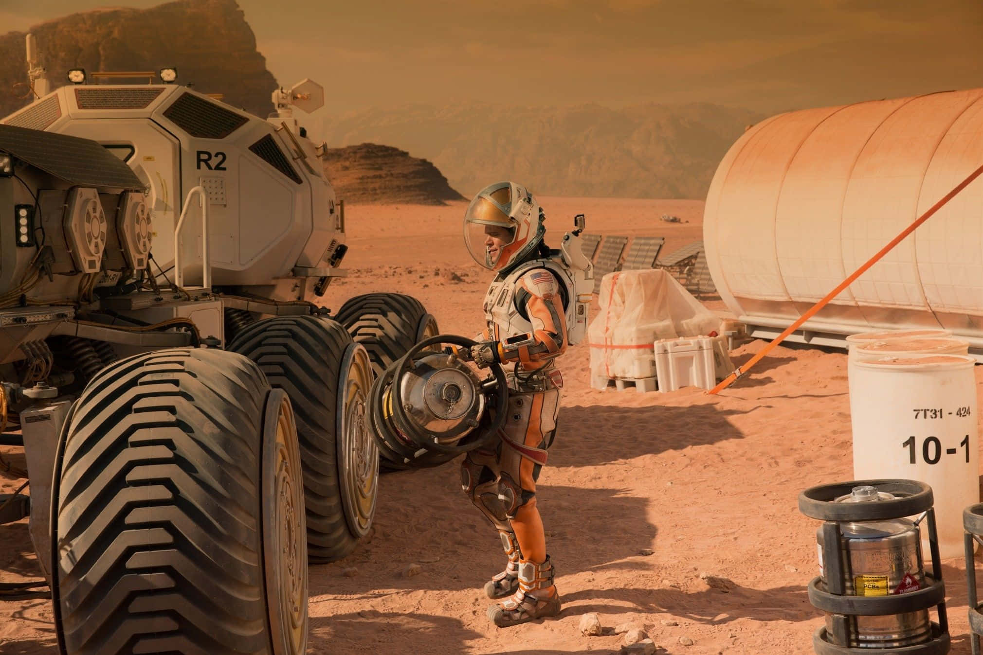 The Martian 桌布 壁紙 — 一名身穿橙色裝備的太空人站在大型探測車旁，背景是佈滿灰塵的紅色火星景觀，並有居住模組。