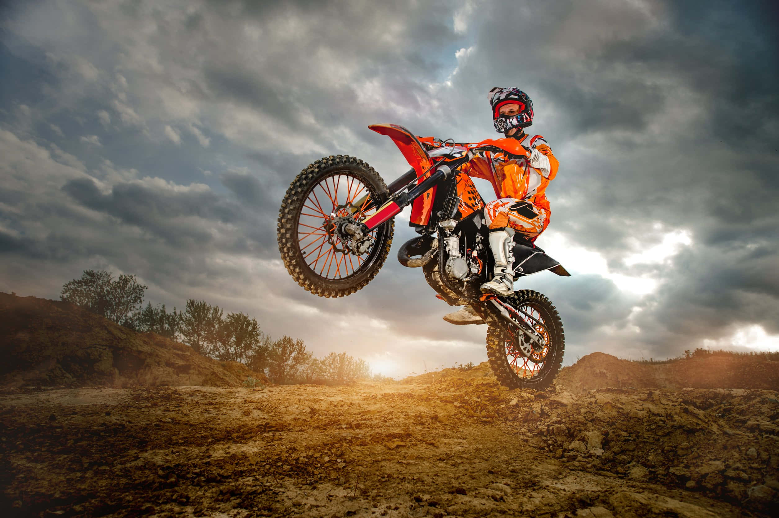 Dirtbike 桌布 壁紙 — 一名身穿橙色裝備的越野摩托車手，在日落時分戲劇性的多雲天空背景下，騎著紅色摩托車在空中飛翔。