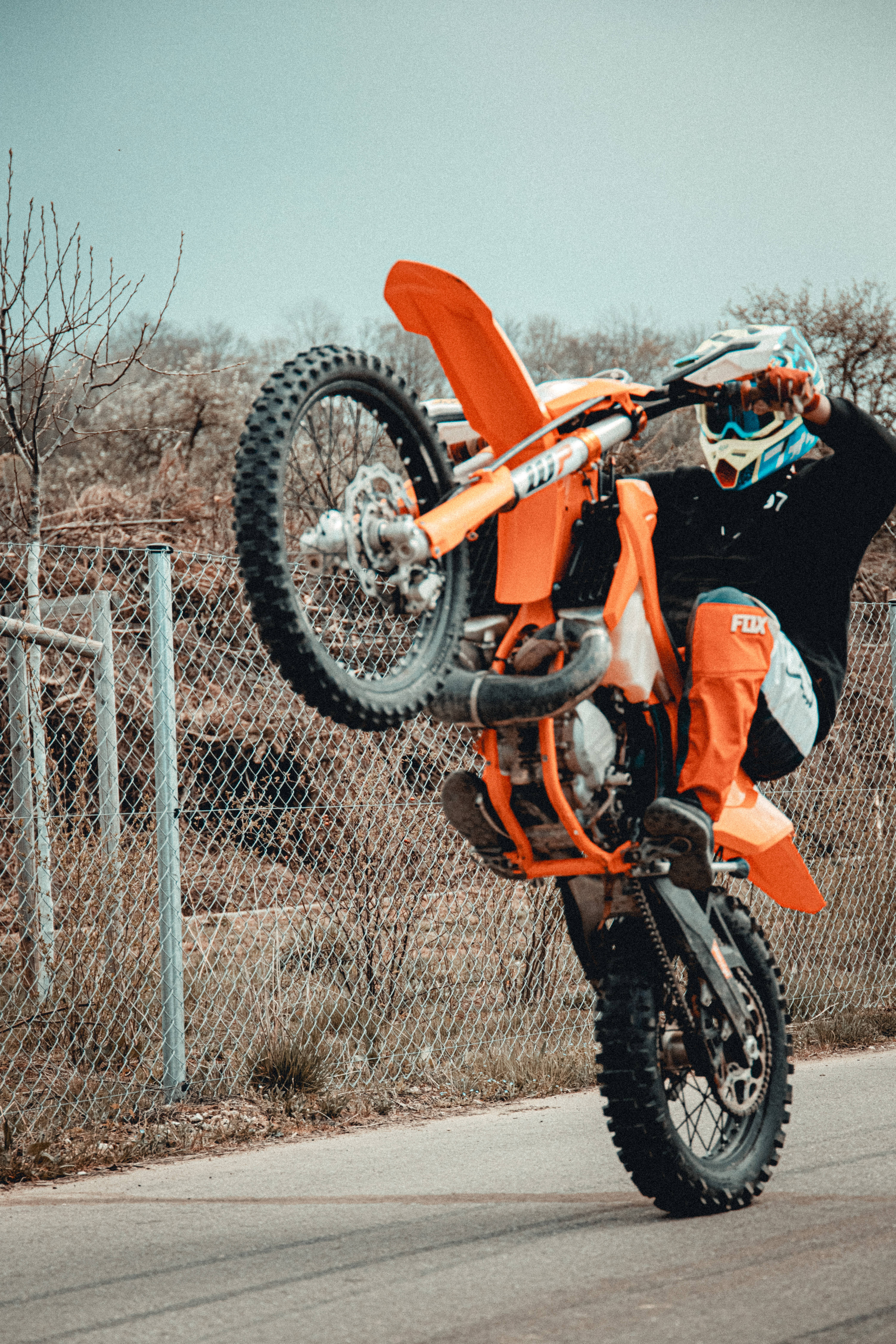 Ktm Bike 桌布 壁紙 — 一名身穿橙色裝備的騎士在鐵絲網圍欄附近，騎著亮橙色的越野摩托車表演翹前輪。