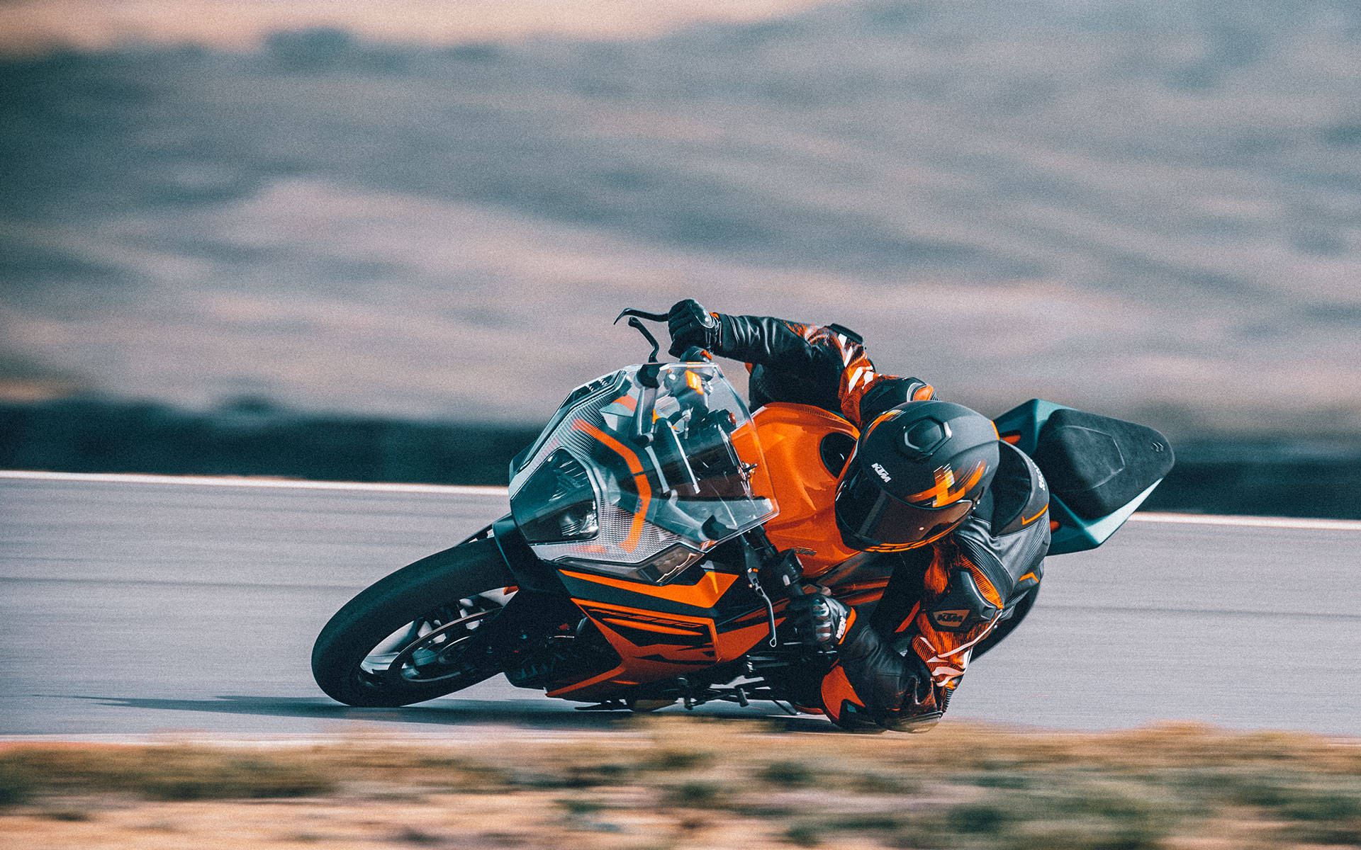 KTM RC 200 桌布 壁紙 — 一名身穿橙色裝備的騎士在陰天下的賽道上，騎著一輛鮮橙色運動機車傾斜入急彎。