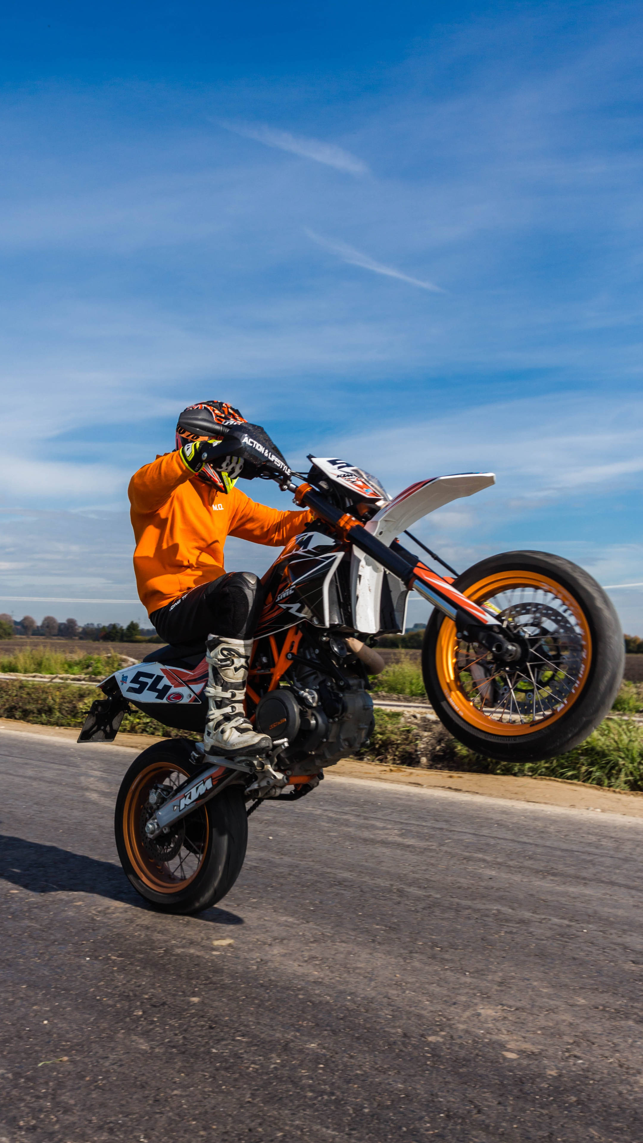 ktm rc 200 桌布 壁紙 — 一名身穿橙色裝備的騎士在陽光明媚的道路上，騎著橙色和白色的KTM越野摩托車表演翹前輪。