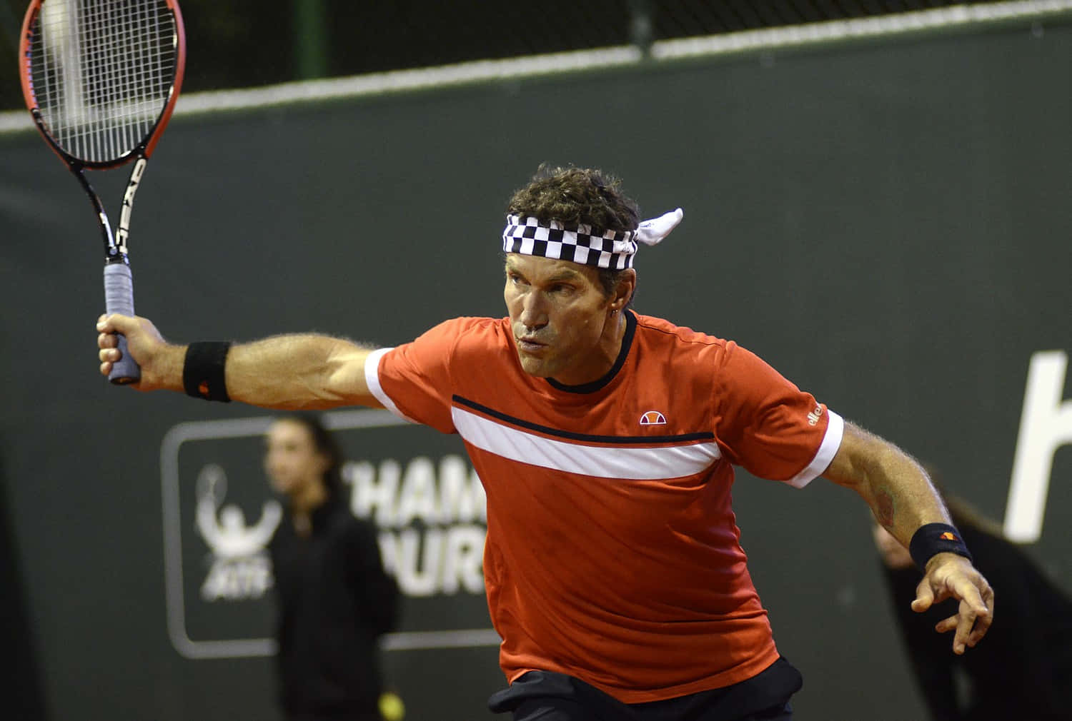 Tennis 桌布 壁紙 — 一名身穿橙色襯衫和格子頭帶的男網球運動員在夜間比賽中專注地揮動球拍。