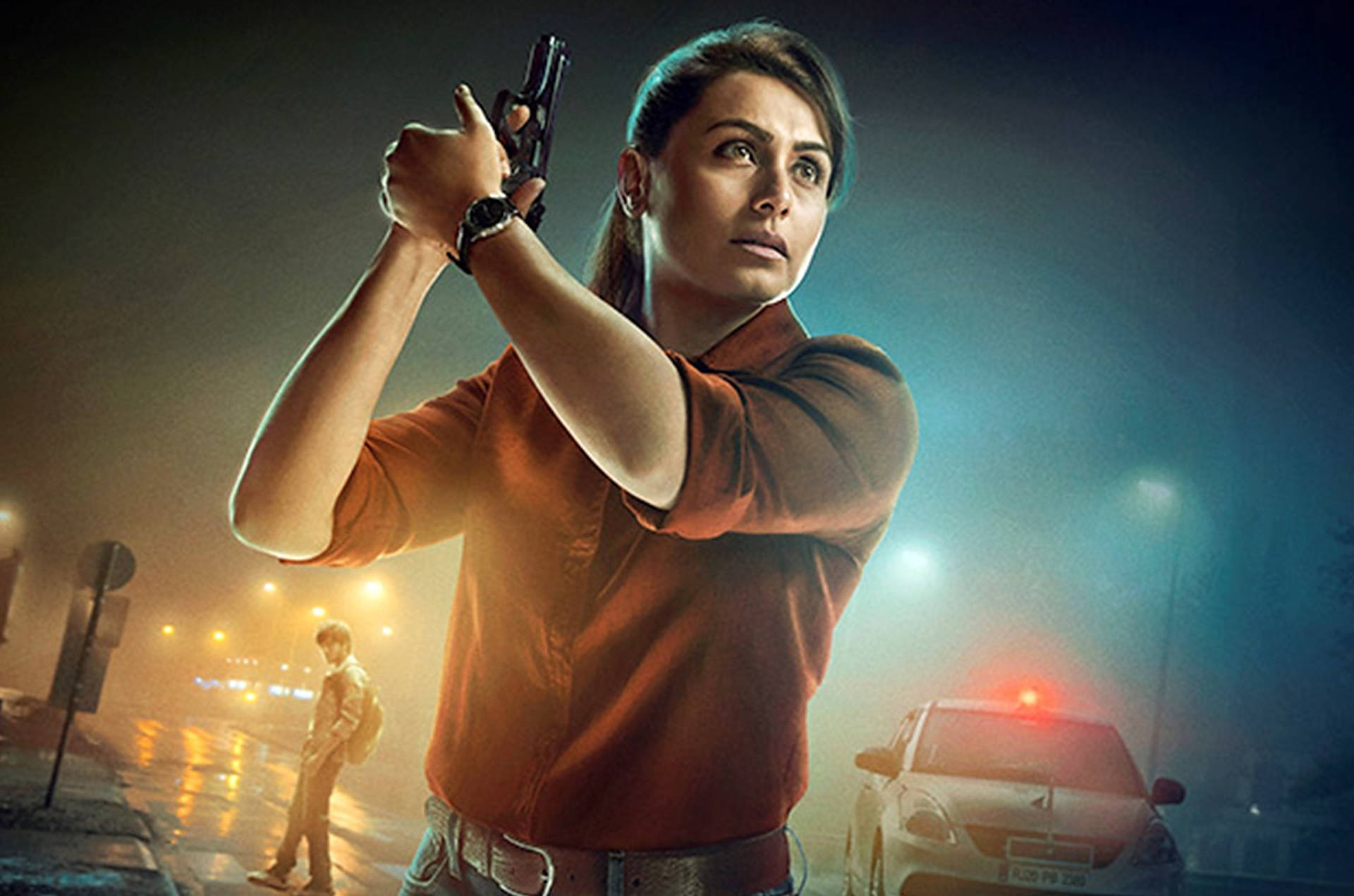 Rani Mukerji 桌布 壁紙 — 一名身穿橙色襯衫的女子在夜晚的霧街上用手槍瞄準，背景有一輛警車和另一個人。