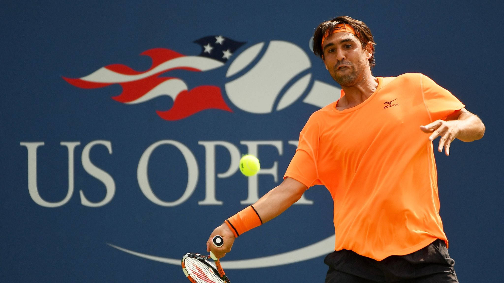 Tennis 桌布 壁紙 — 一名身穿橙色襯衫的男性網球選手在藍色美國網球公開賽背景下揮拍擊打黃色網球。