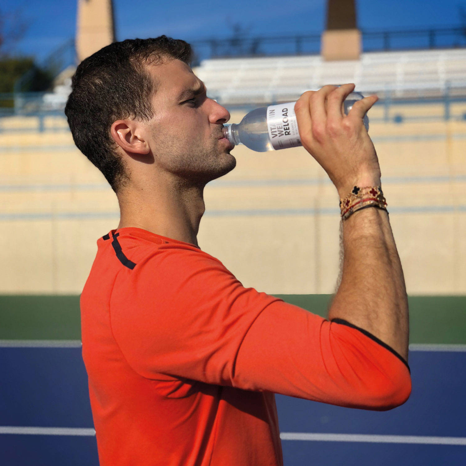 Drink Water 桌布 壁紙 — 一名身穿橙色襯衫的男性運動員在陽光明媚的網球場上喝水，背景有觀眾看台。