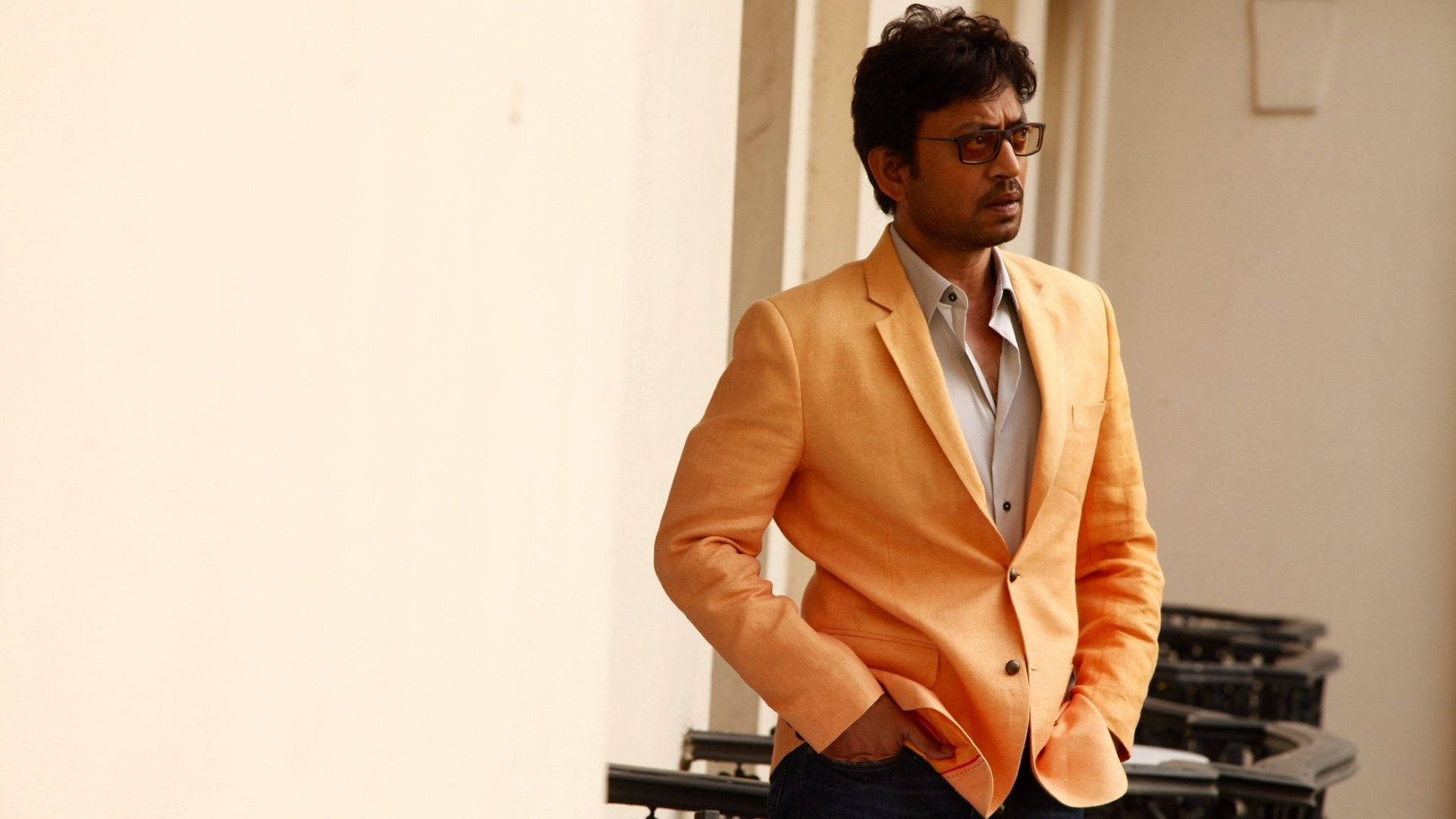Irrfan Khan 桌布 壁紙 — 一名身穿橙色西裝外套、戴眼鏡的男子，雙手插袋，自信地站在純色米色牆前。