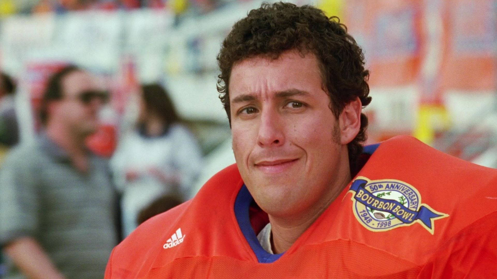 Adam Sandler 桌布 壁紙 — 一名身穿橙色足球球衣、帶有50週年紀念徽章的男人尷尬地對著鏡頭微笑。