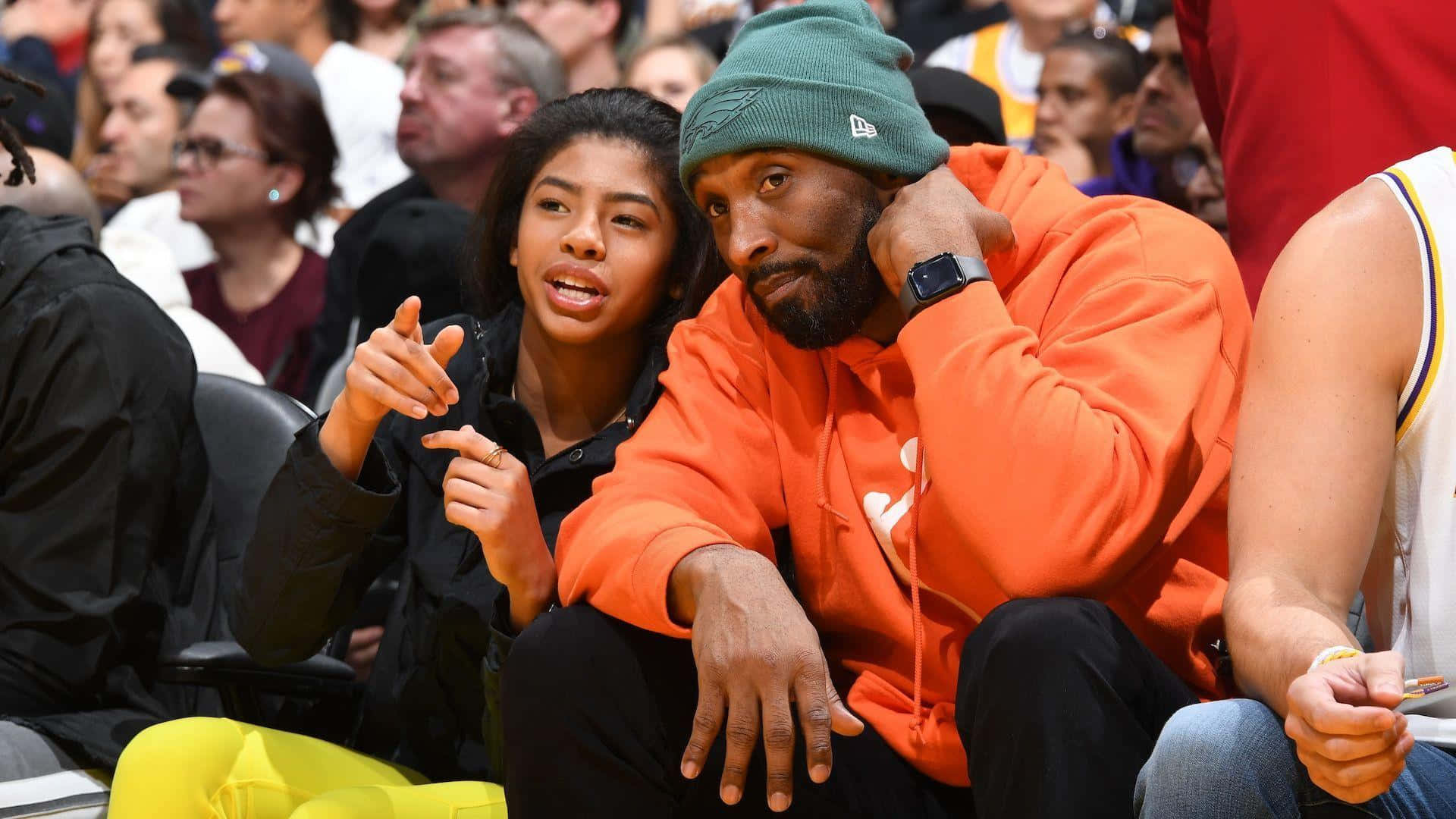 Kobe And Gigi 桌布 壁紙 — 一名身穿橙色連帽衫和綠色毛帽的男子坐在一名身穿黑色外套的年輕女子旁邊，兩人都專注地看着擁擠場館內的籃球比賽。