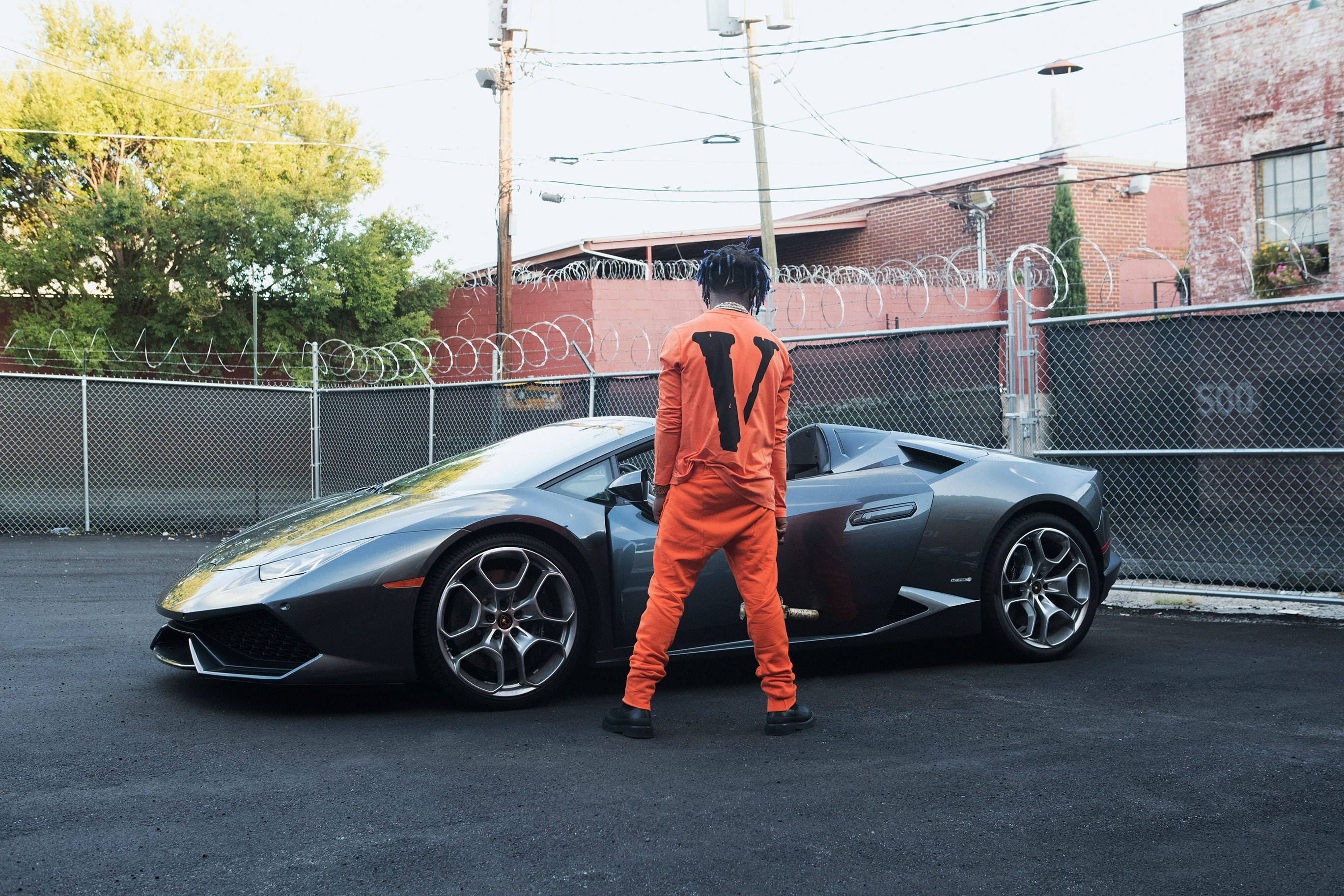Vlone 桌布 壁紙 — 一名身穿橙色連身衣的人站在一個有鐵絲網圍欄和磚砌建築的市區空地，旁邊是一輛時尚的灰色藍寶堅尼。