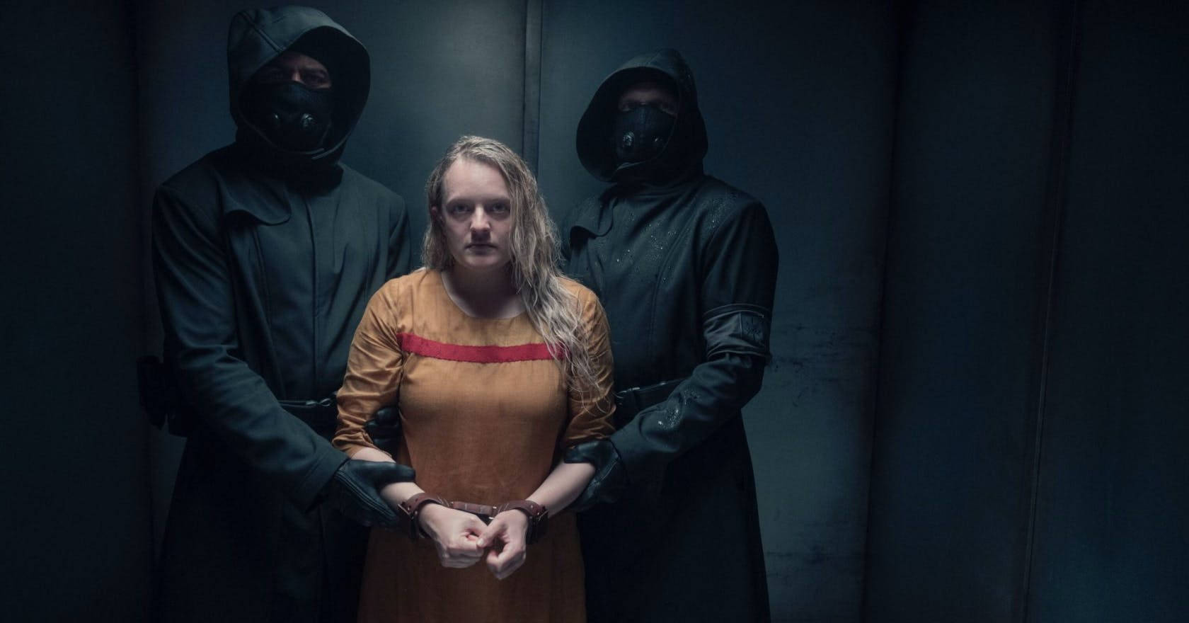 The Handmaid's Tale 桌布 壁紙 — 一名身穿橙色連身衣的女子，在燈光昏暗的審訊室內，被戴上手銬，站在兩名戴頭套面具的警衛之間。